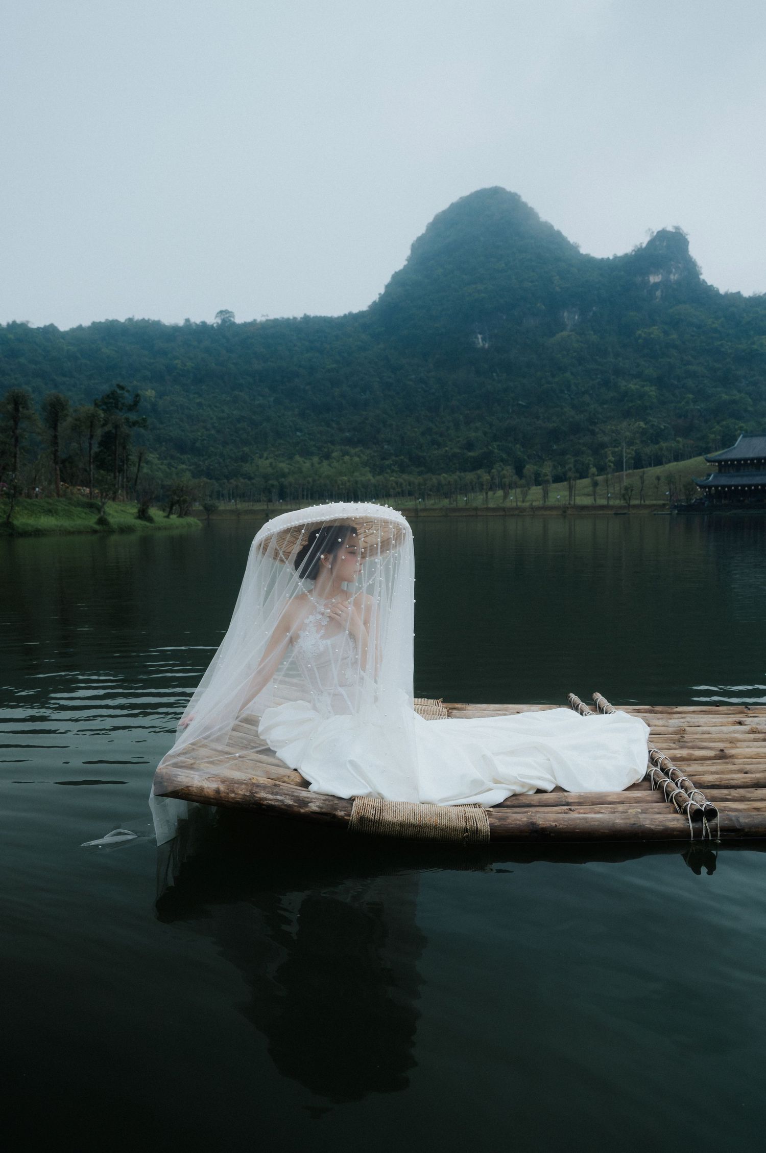 Bride in white gown and veil resting on bamboo raft on calm lake with lush green karst mountains backdrop.