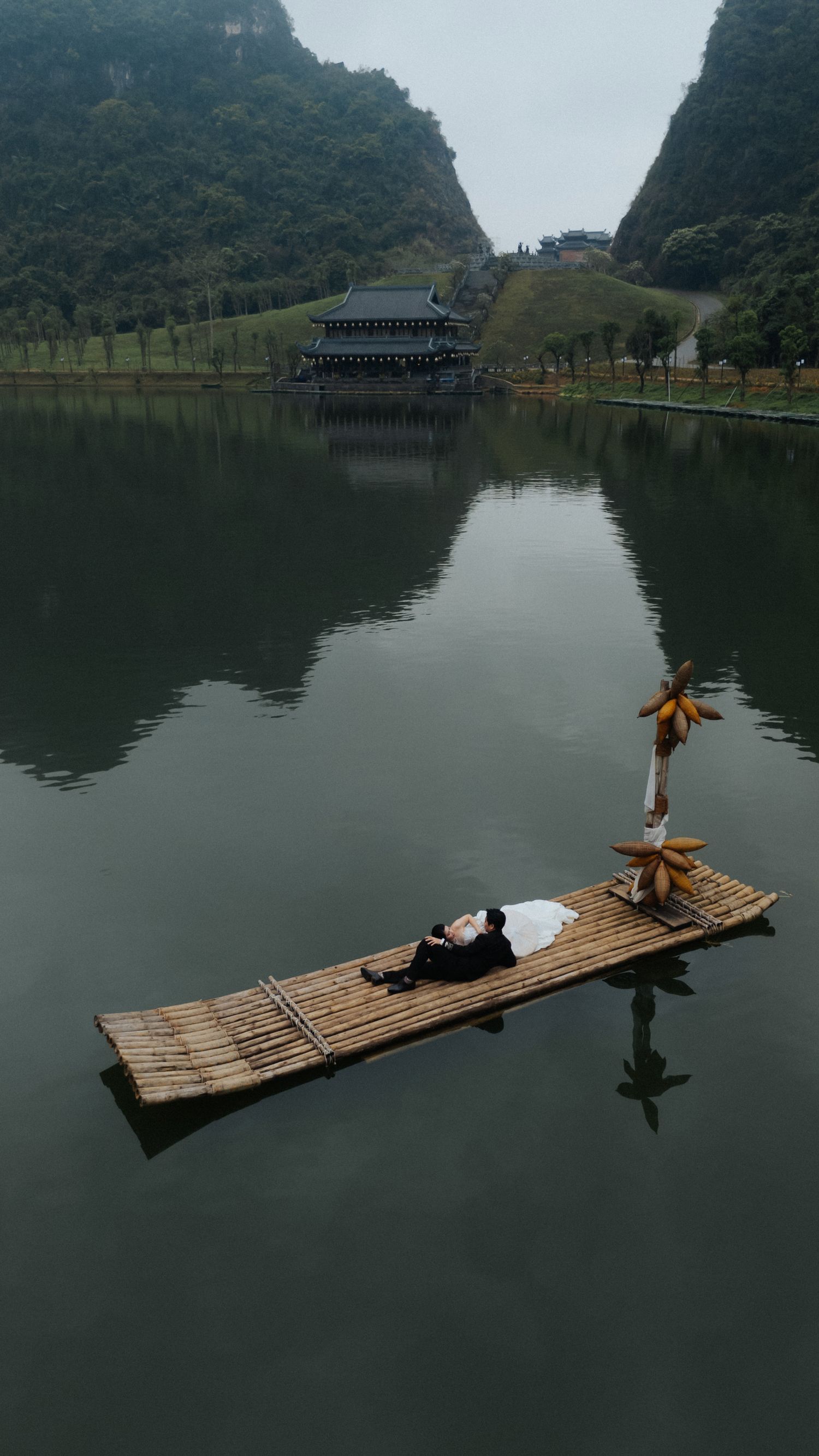 Person lying on bamboo raft floating on calm lake with karst mountains and traditional Chinese pavilion in background.