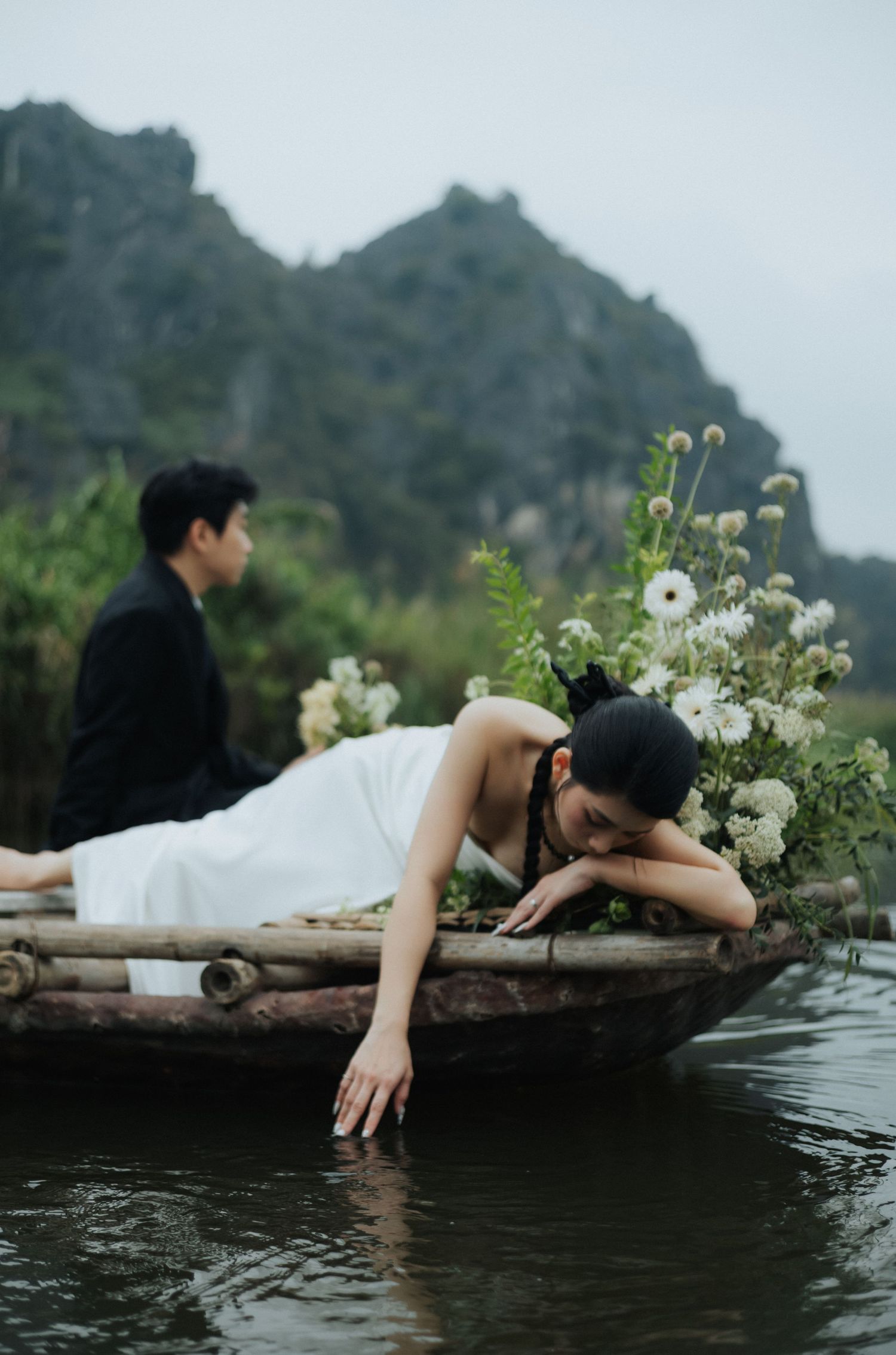 A woman in a white dress leans over a wooden boat touching water, with a man in black and misty mountains behind her.