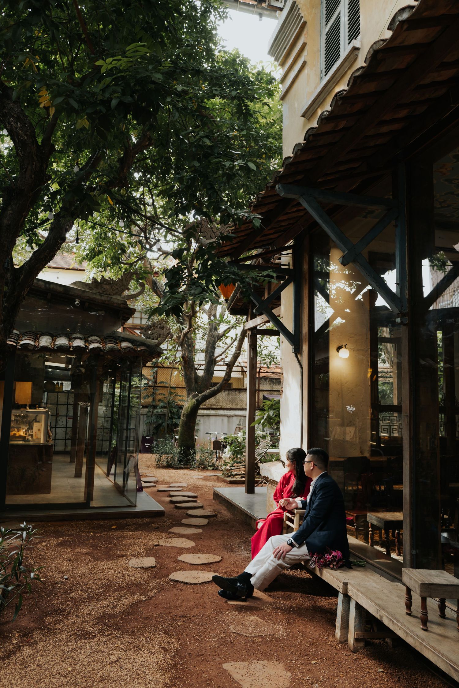 Couple sitting together on a wooden porch in a lush courtyard setting, surrounded by trees and traditional architecture.