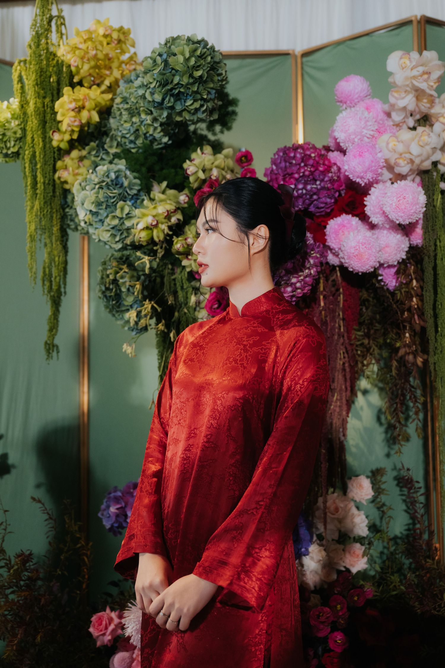 Young woman in red Vietnamese ao dai dress stands before a decorative floral backdrop with colorful blooms.