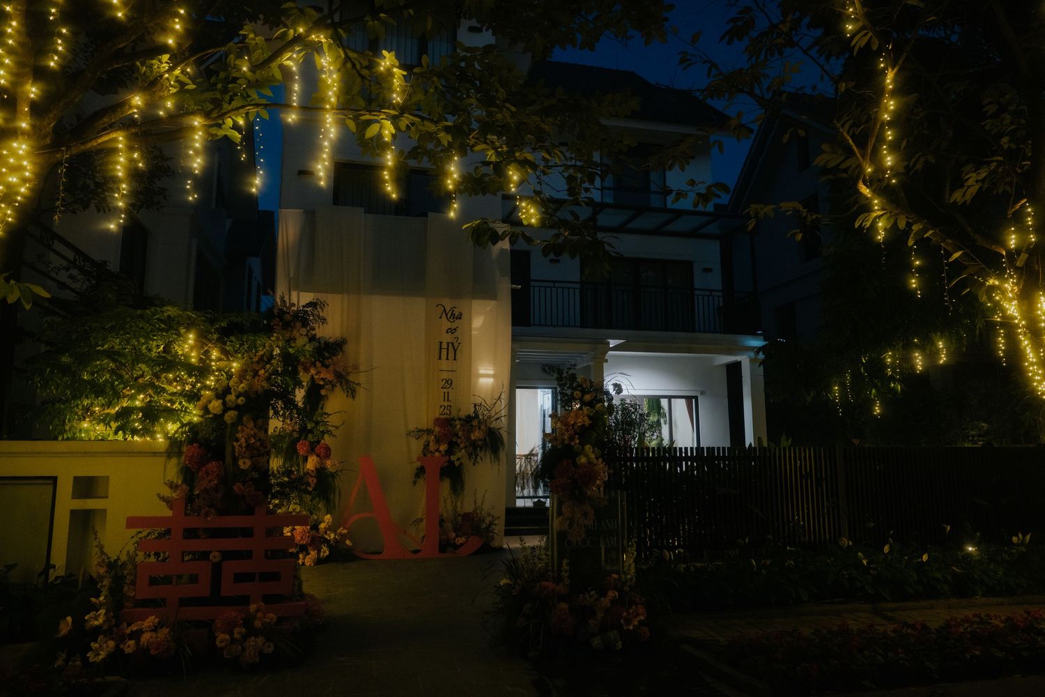A beautifully decorated house at night with warm fairy lights hanging from trees and floral arrangements at the entrance.