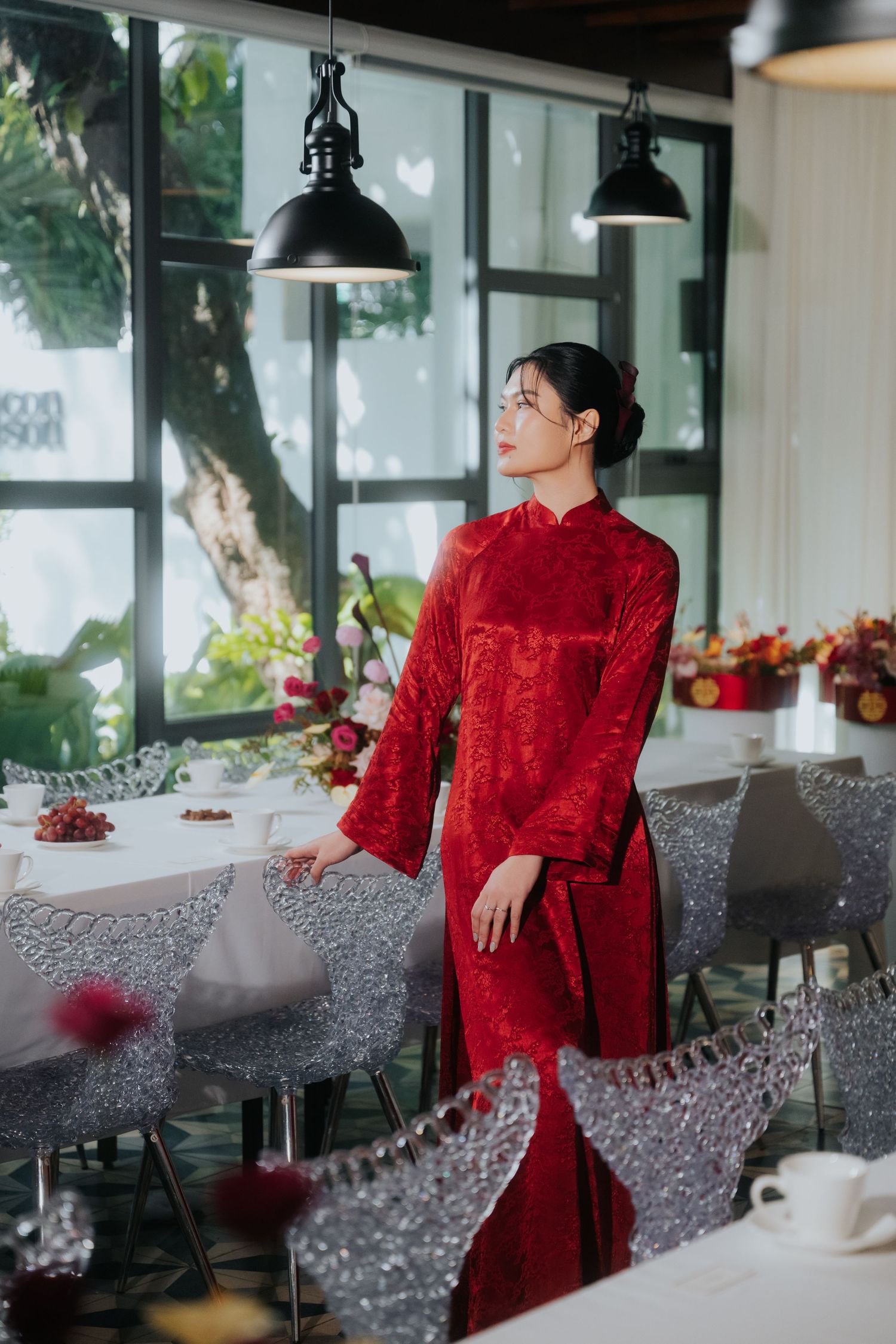 A woman in a red Vietnamese ao dai dress stands elegantly in a modern restaurant with crystal chairs and floral decor.