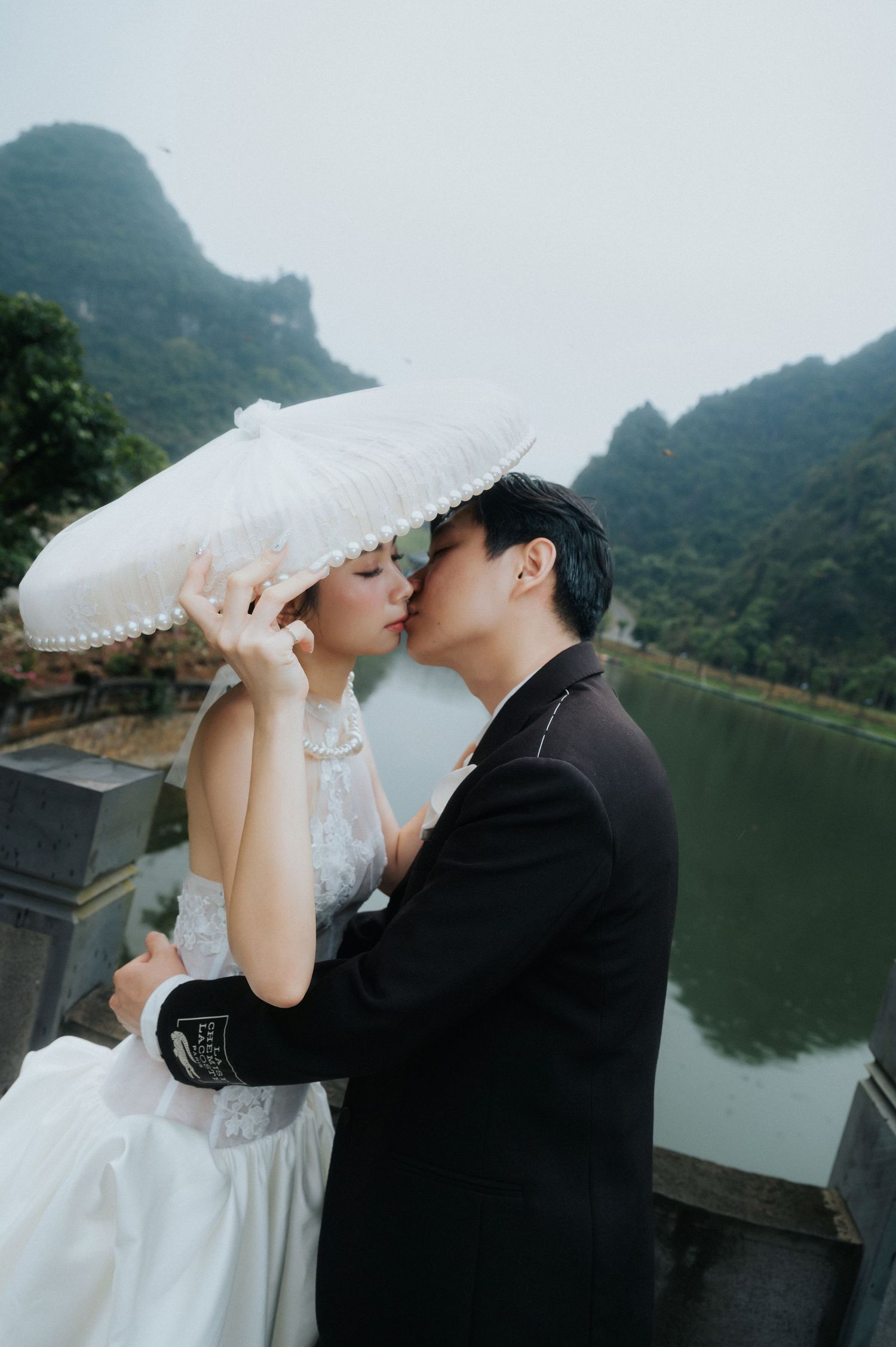 A couple shares a romantic kiss under a white parasol in wedding attire, with misty mountains and a river in the background.