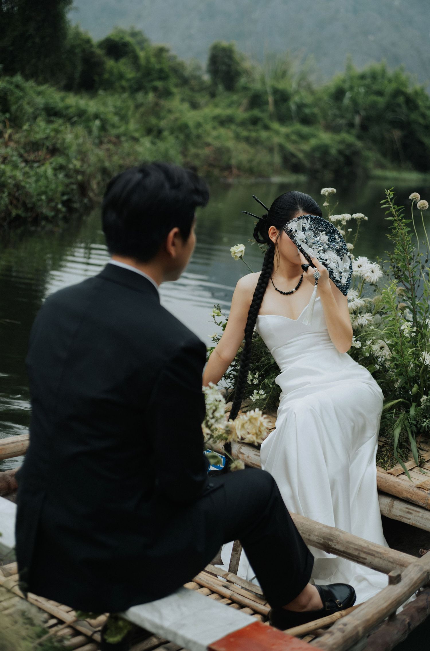 A couple in wedding attire sits on a wooden boat surrounded by flowers, with a serene river and lush greenery in the background.