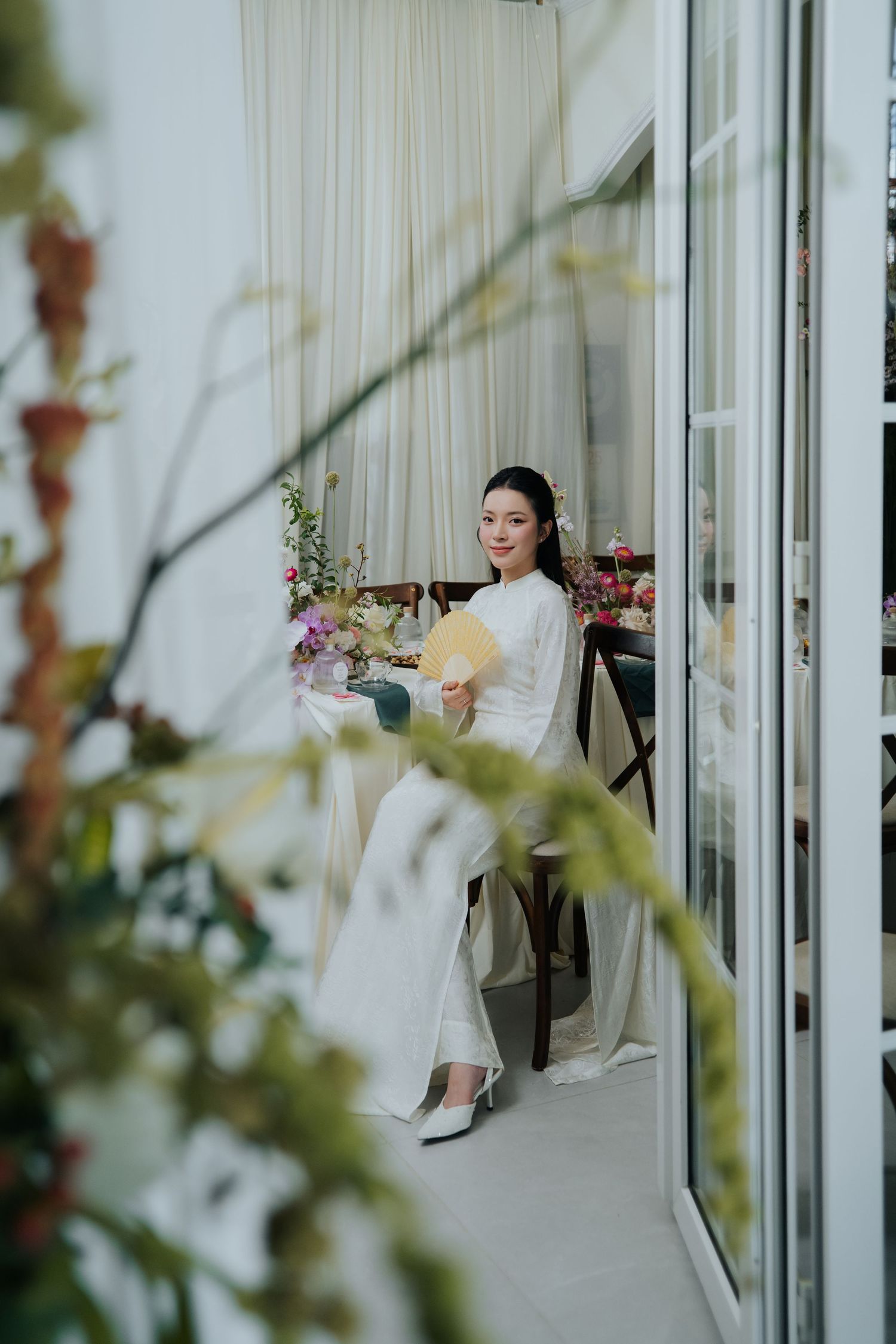 A bride in a white ao dai holds a yellow fan, seated gracefully among flowers in an elegant, light-filled room.
