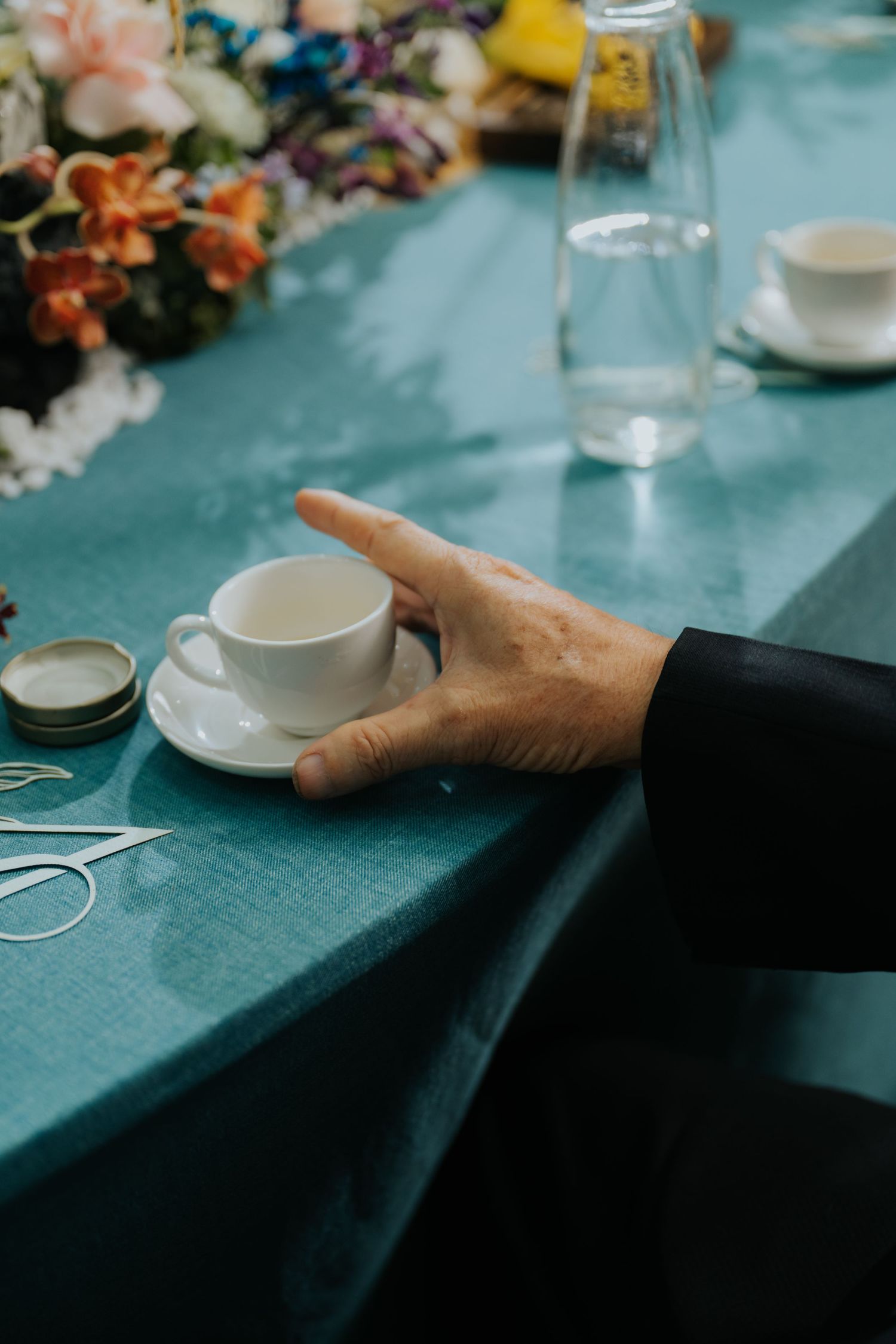 Hand reaching for a white espresso cup on a teal tablecloth with flowers and glassware at an elegant event.