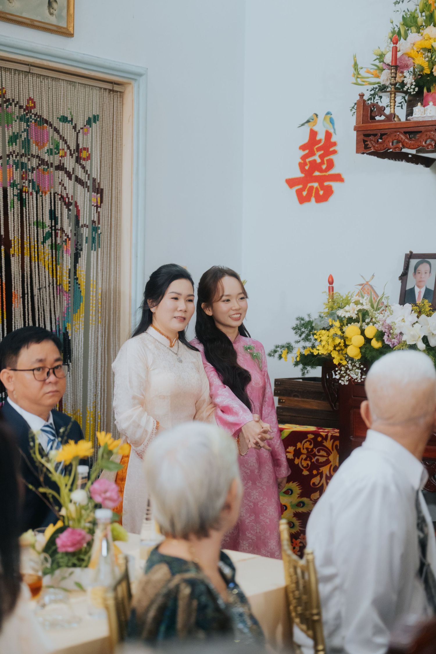 Two women in traditional Vietnamese ao dai stand at a wedding ceremony with double happiness symbol on wall.