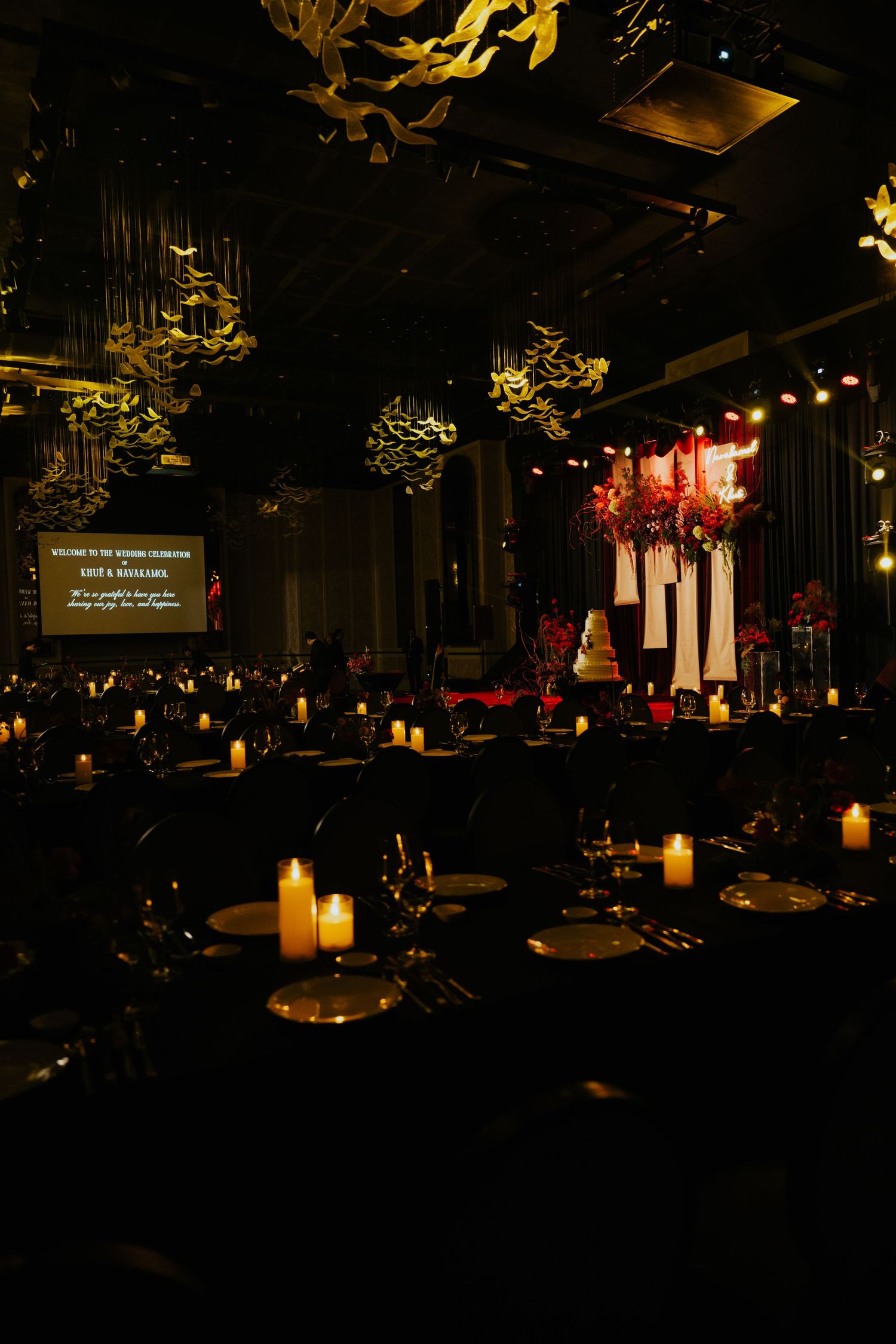 Elegant dark wedding reception hall with candlelit tables, dramatic floral installations, and a glowing projection screen.