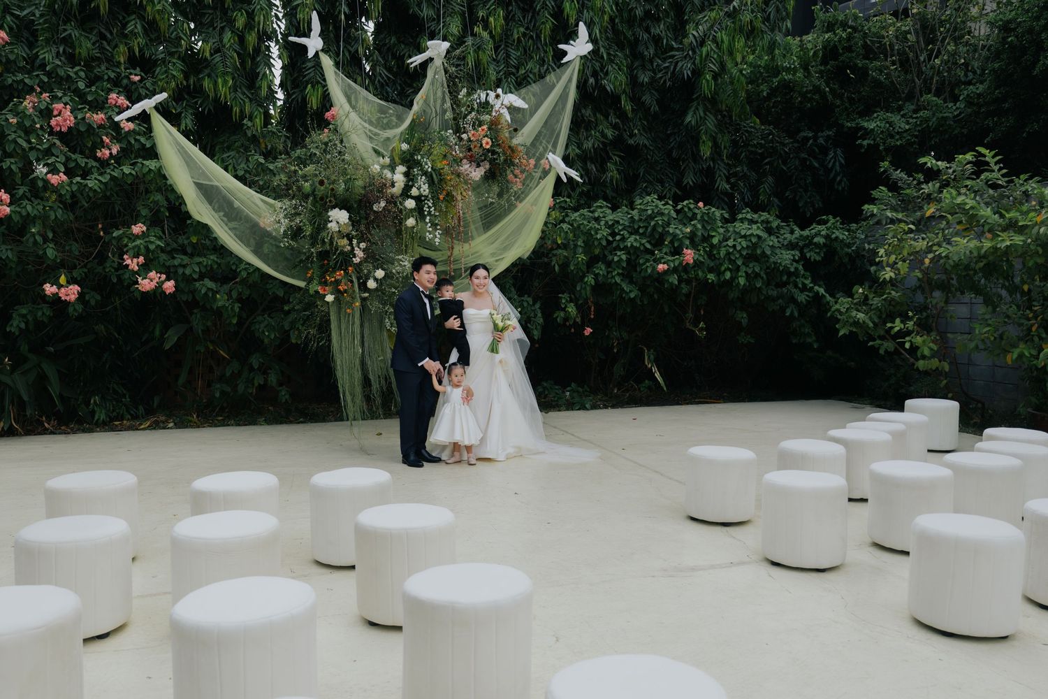 Bride and groom pose at an elegant outdoor wedding ceremony with white ottomans and a floral arch backdrop.