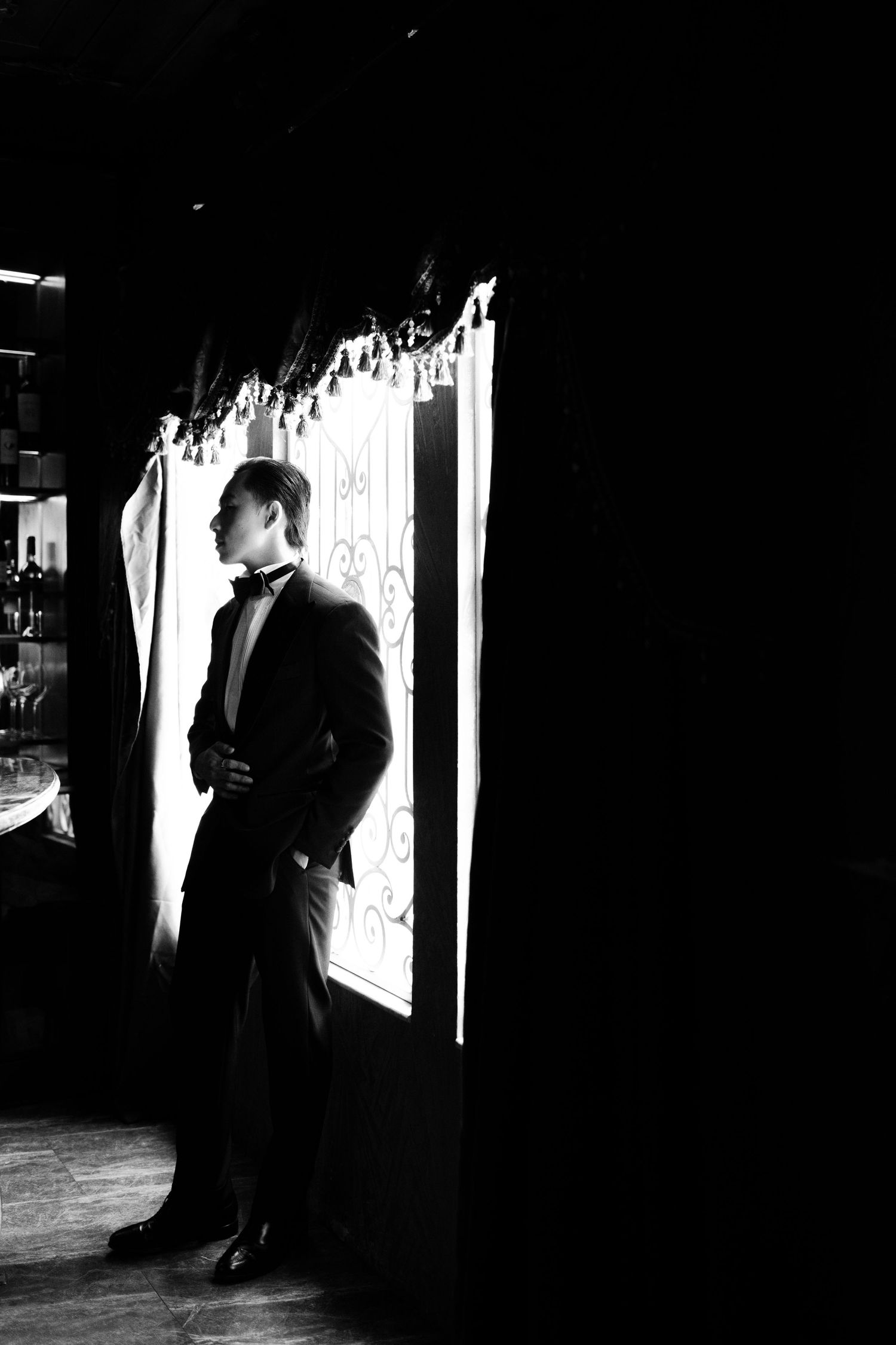 Black and white photo of an elegant man in a tuxedo standing by a bright window in a dark, sophisticated interior.