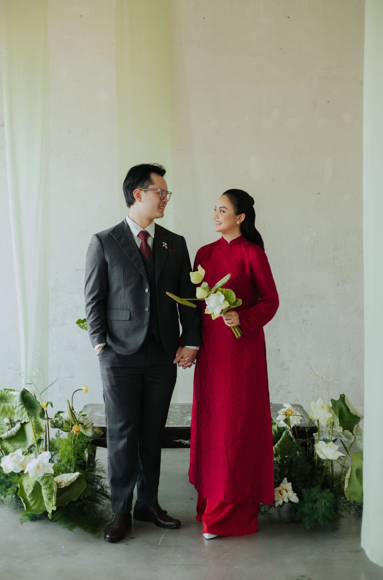 Couple in traditional Vietnamese ao dai and suit holding hands with white flowers and green foliage backdrop.