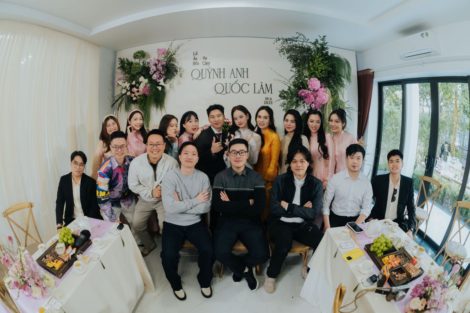 Group photo of wedding party guests posing indoors with floral decorations and a sign reading Quynh Anh Quoc Lam.