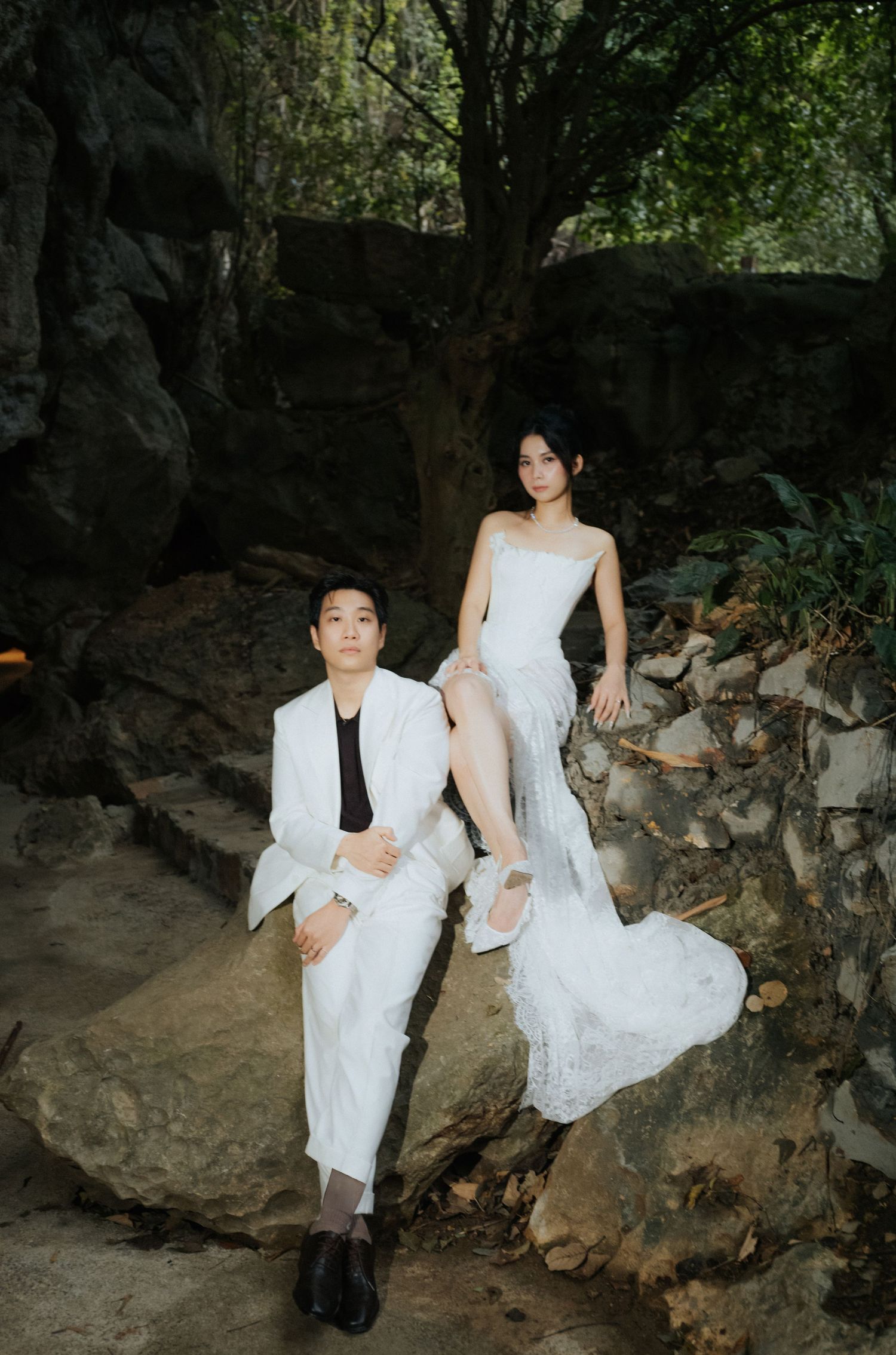 A couple in white outfits poses on rocky steps surrounded by lush greenery, evoking an elegant pre-wedding photo shoot.