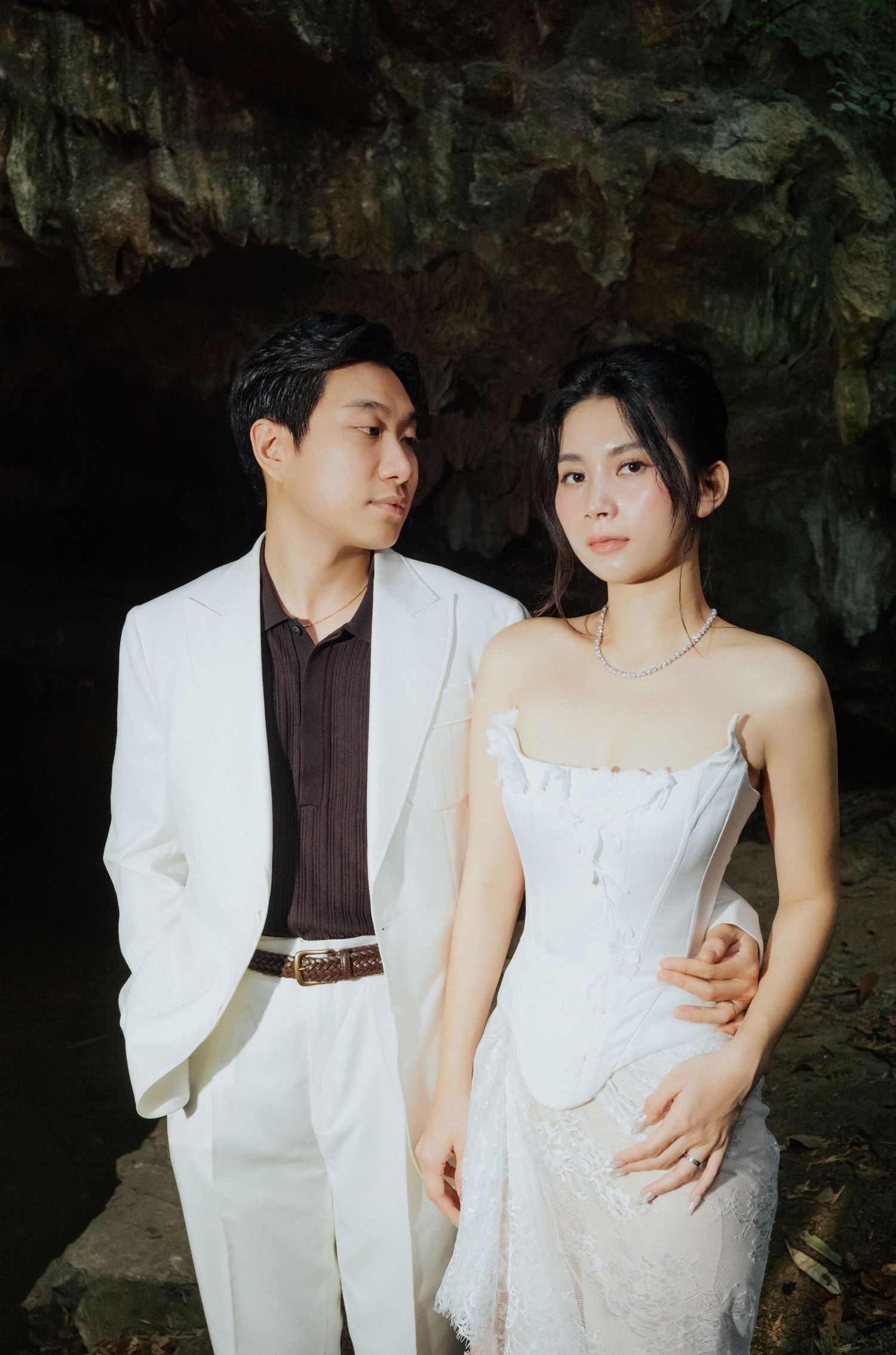 A couple in white wedding attire poses romantically inside a cave, the man in a suit and woman in a strapless lace dress.
