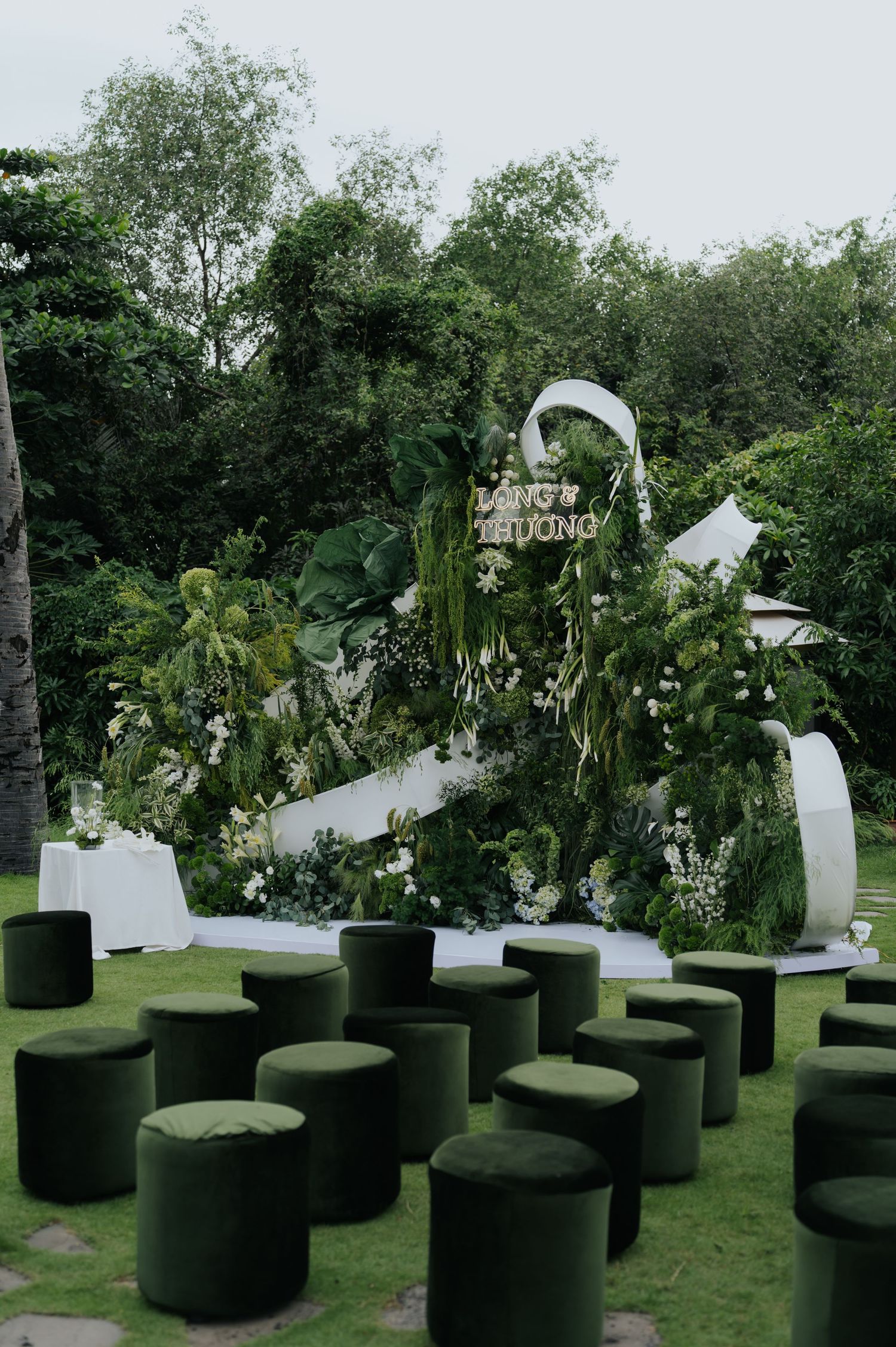 Outdoor wedding ceremony setup with lush greenery backdrop, white floral arch, and rows of dark green velvet ottoman seats on grass.