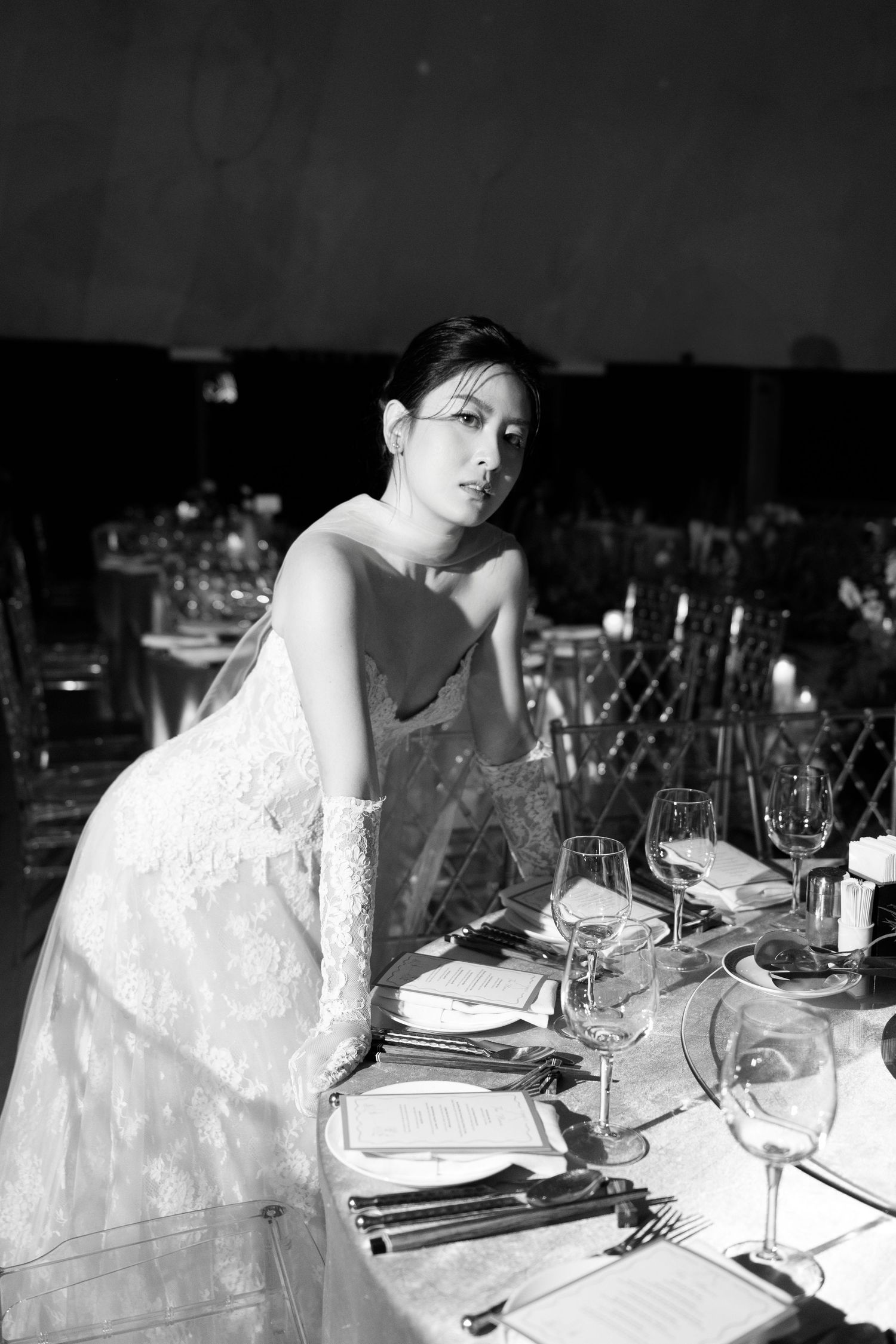 Black and white photo of a bride in lace gown seated at an elegantly set wedding reception table with wine glasses.