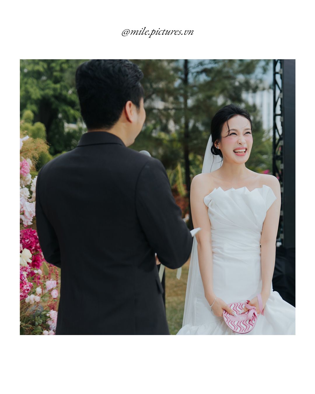 A bride in a white strapless gown smiles joyfully at her groom in a black suit during an outdoor wedding ceremony.