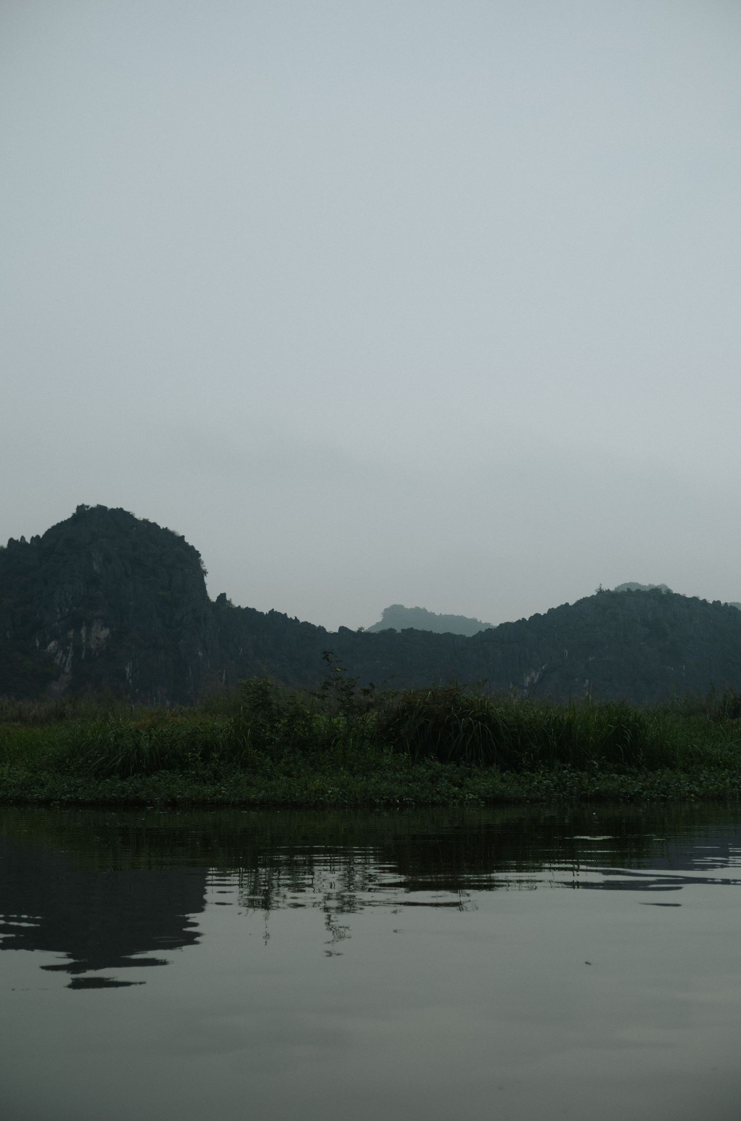 Misty karst mountains reflected in calm river waters with lush green vegetation along the banks in Vietnam.