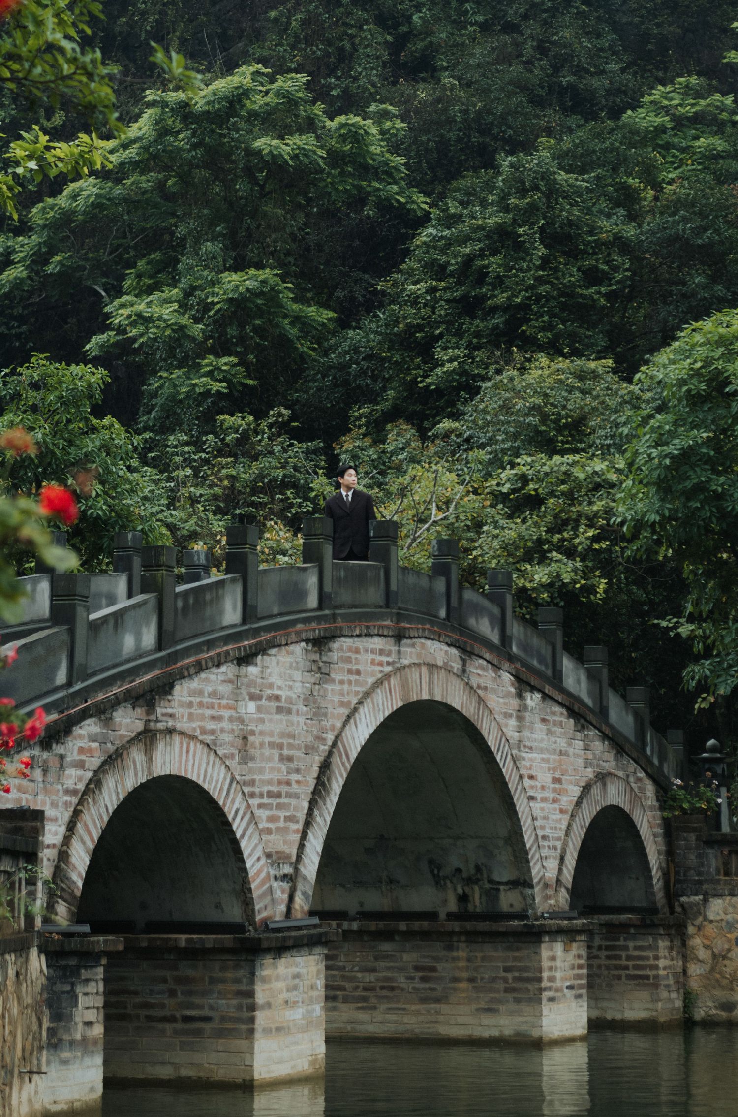 Person standing on a historic stone arch bridge over water surrounded by lush green trees in a serene park setting.