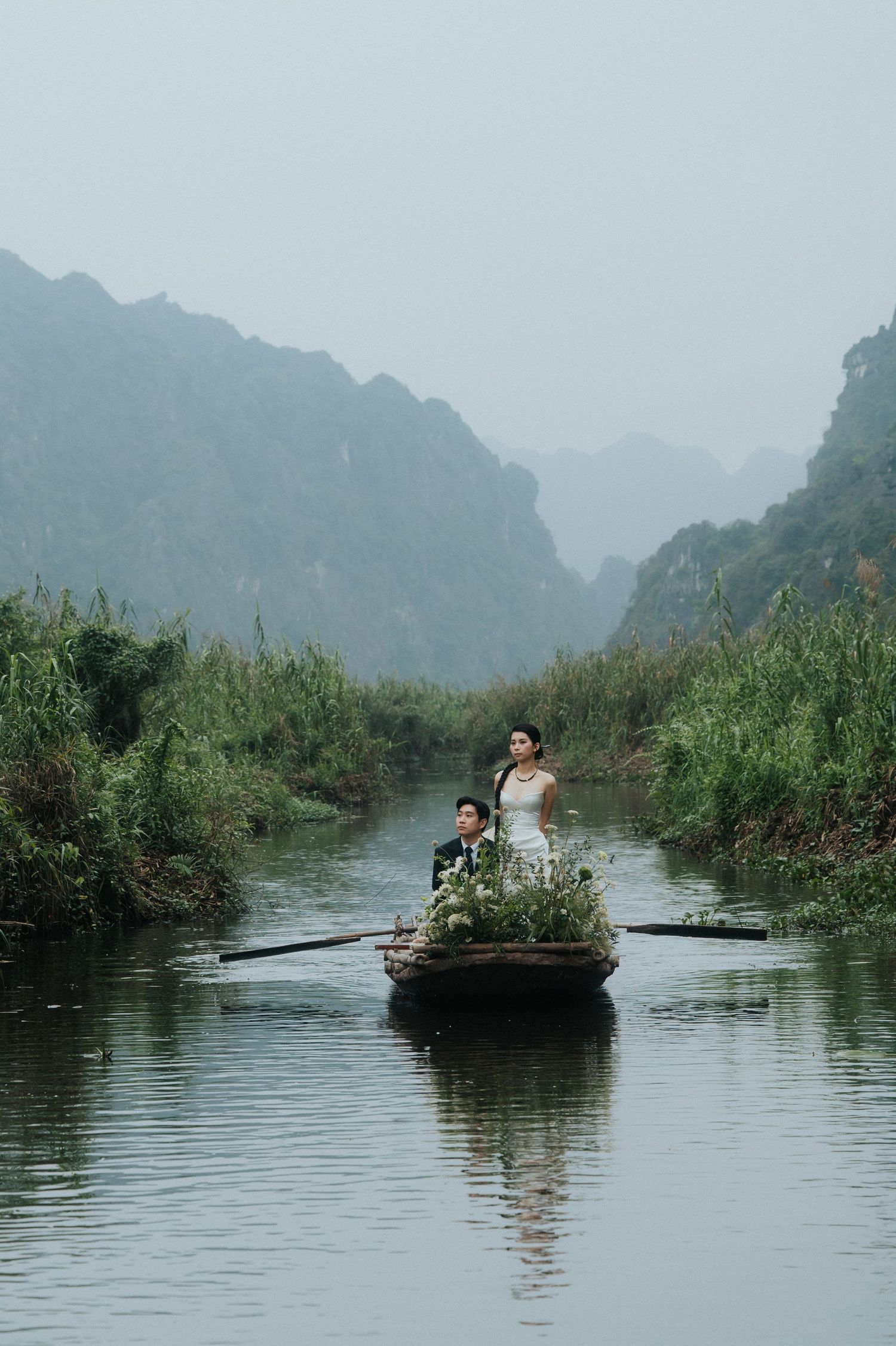 Couple in wedding attire on a rustic boat with flowers, gliding through a misty river surrounded by lush greenery and karst mountains.