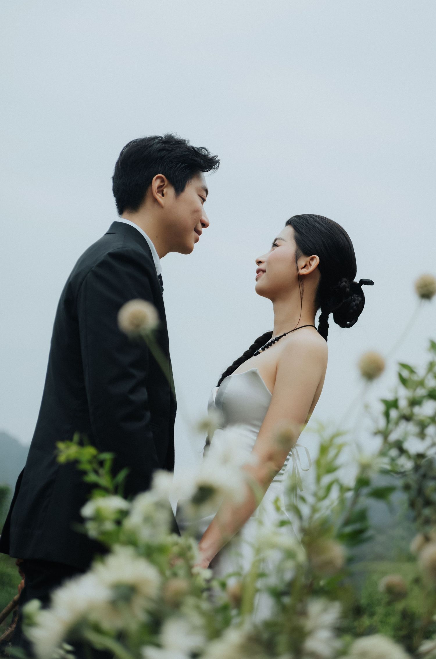 A couple in wedding attire shares a romantic moment surrounded by wildflowers against a misty sky.