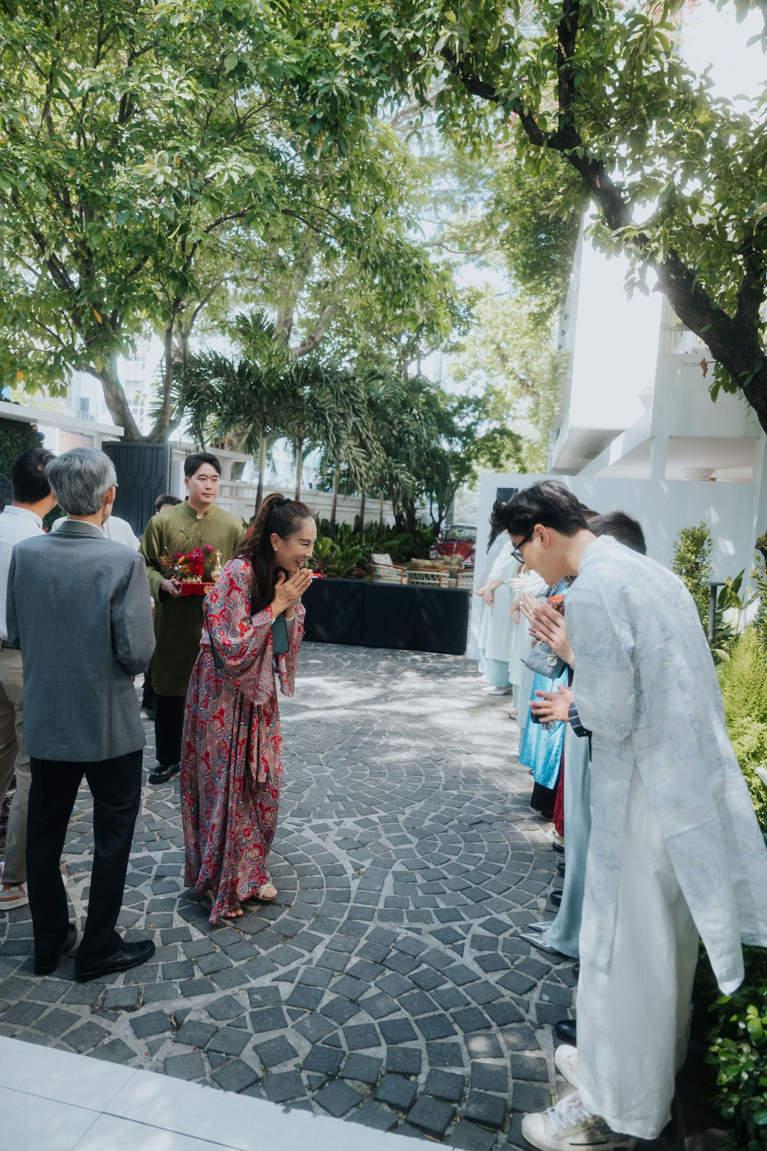 Guests in traditional Vietnamese ao dai attire greet each other at an outdoor wedding ceremony surrounded by lush trees.