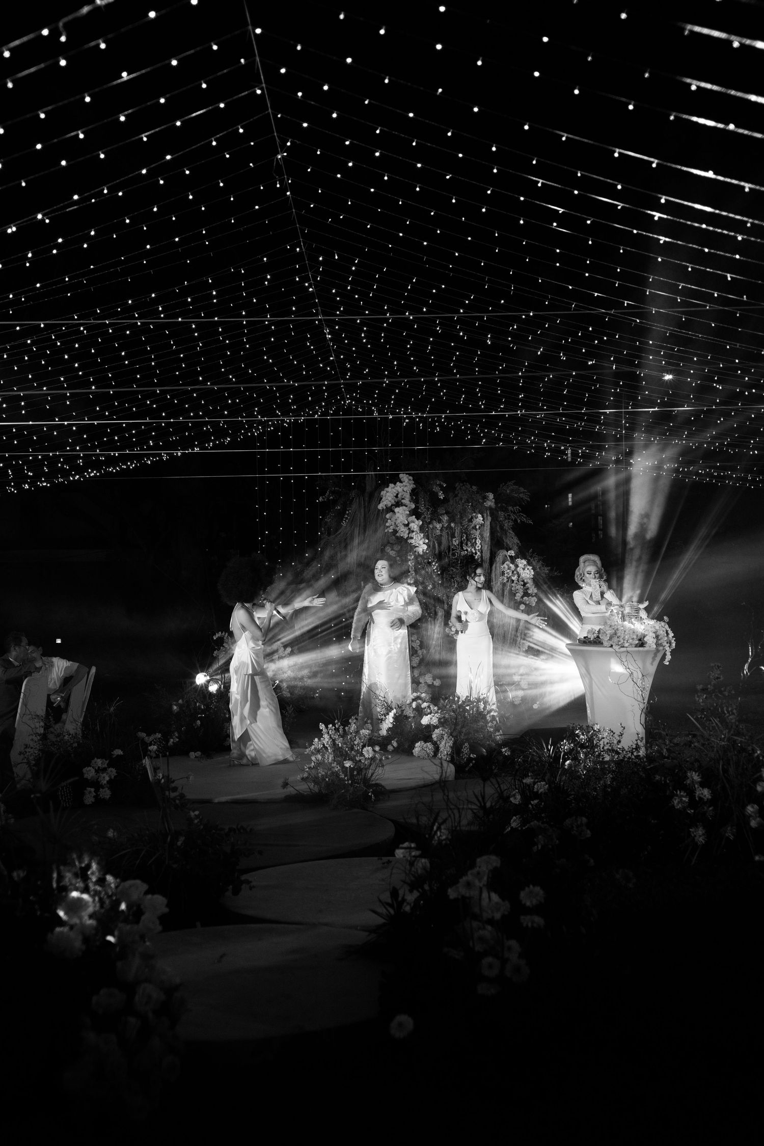 Black and white wedding ceremony under twinkling lights with bridal party on decorated stage.