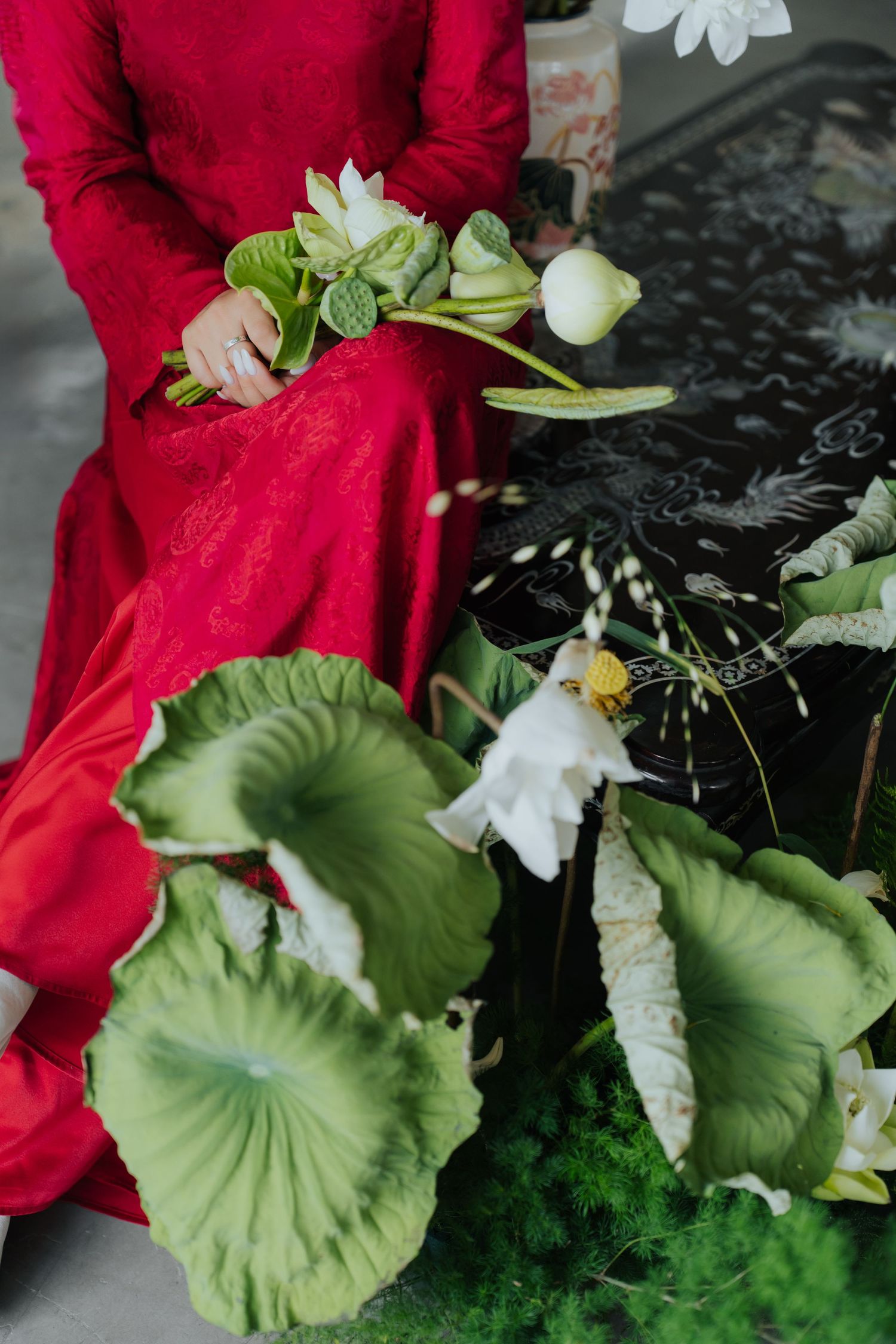 Woman in red ao dai holding white lotus flowers beside large green lotus leaves.