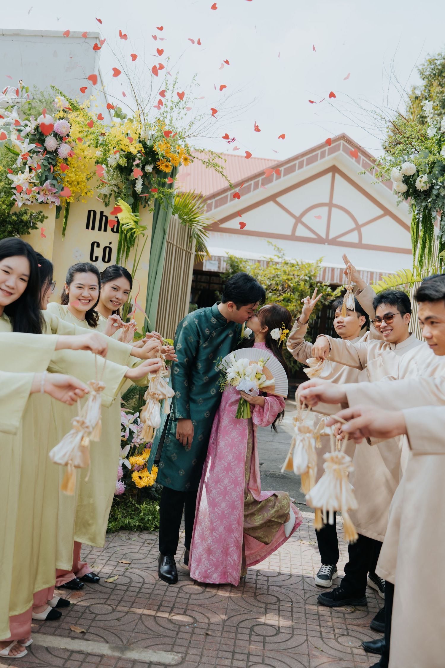 Vietnamese couple kisses during traditional wedding ceremony while guests toss red heart confetti outdoors.