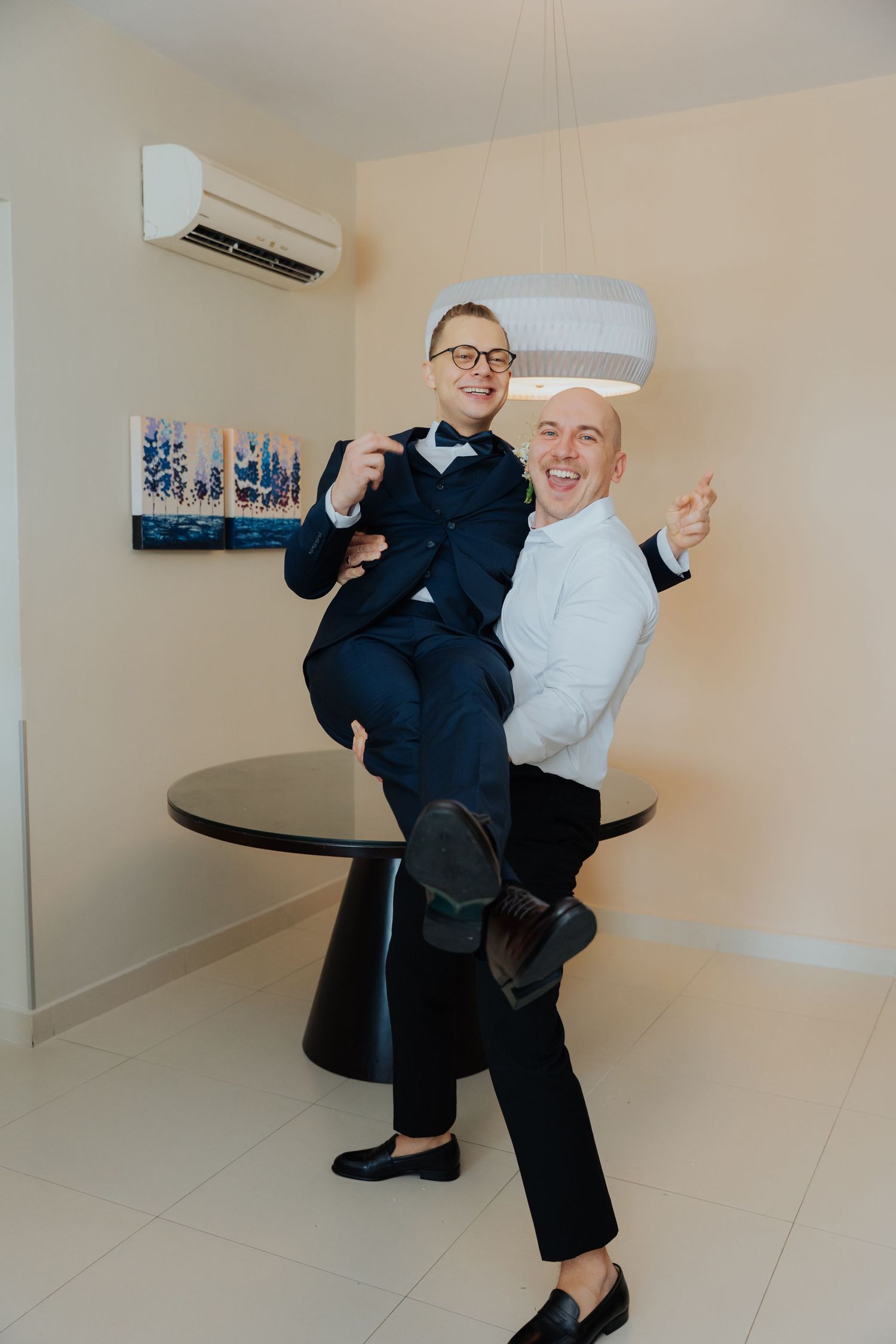Two men in formal suits celebrating joyfully, one lifting the other in a bright modern room with air conditioning.