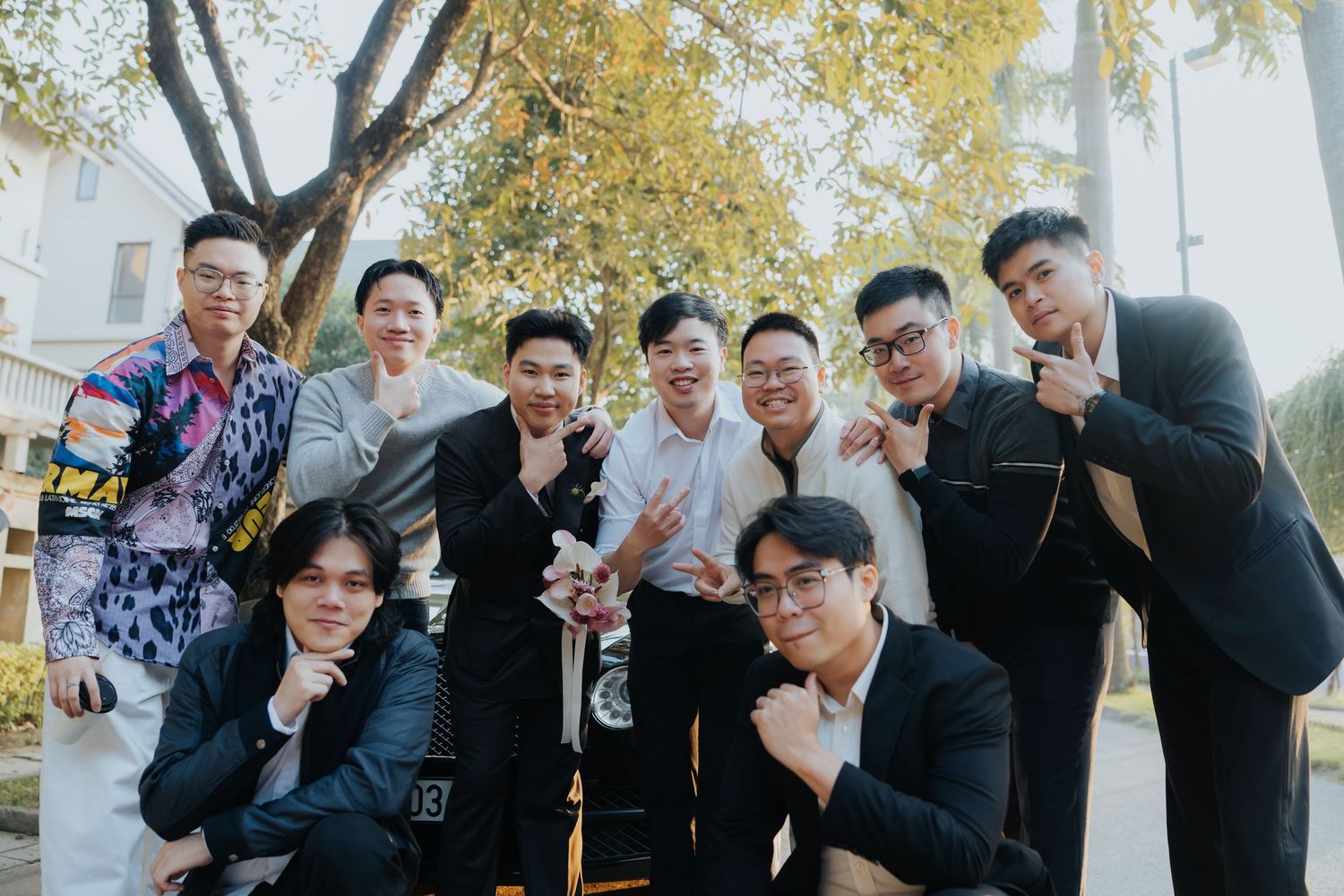 Group of nine young men posing together outdoors under a yellow tree, dressed in formal and casual attire.