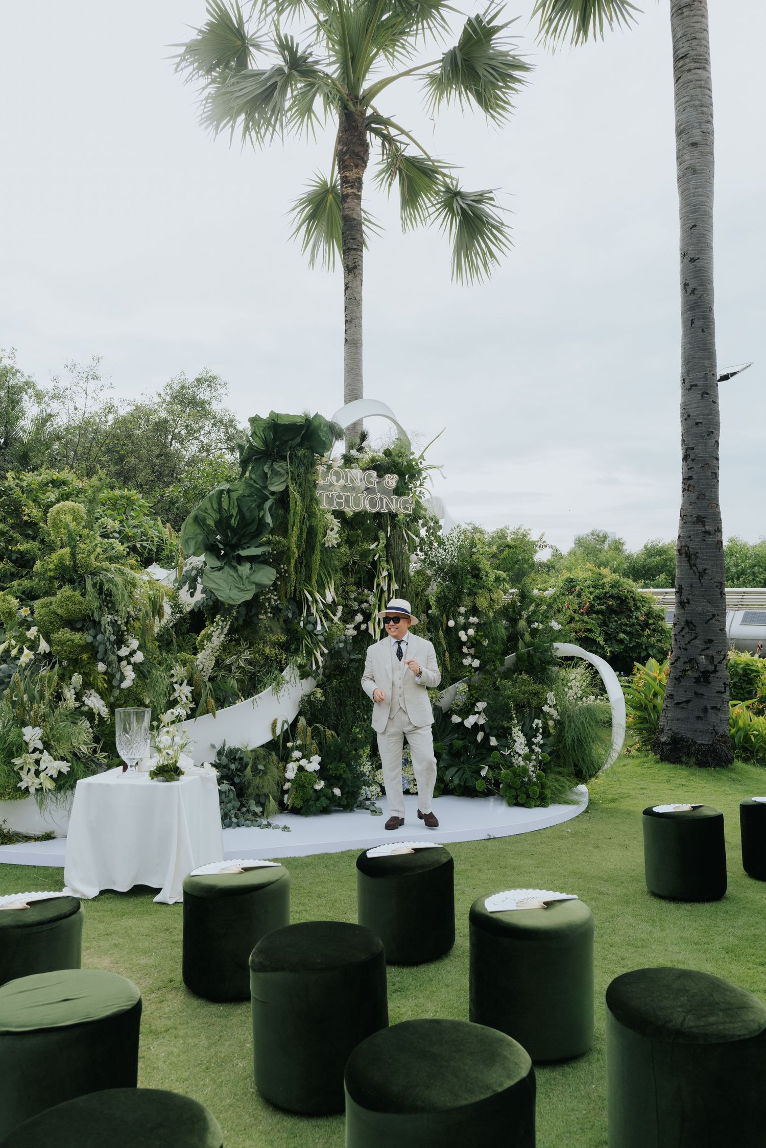 Outdoor wedding ceremony with lush greenery backdrop, palm trees, green velvet ottomans, and officiant on white stage.