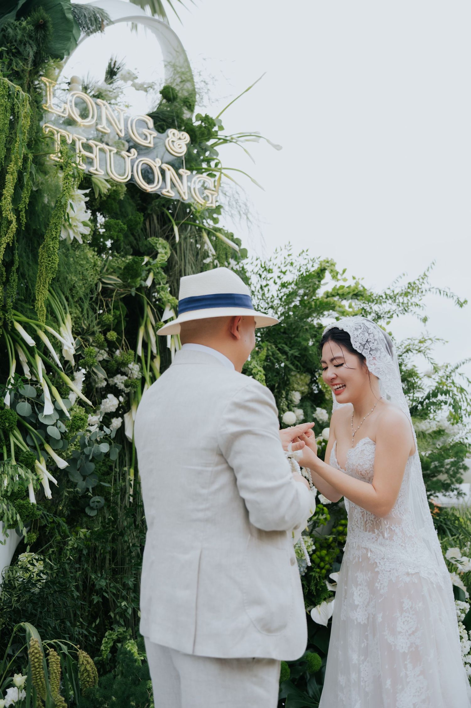 Groom in white suit and hat exchanges rings with smiling bride in lace gown at lush green floral wedding altar.