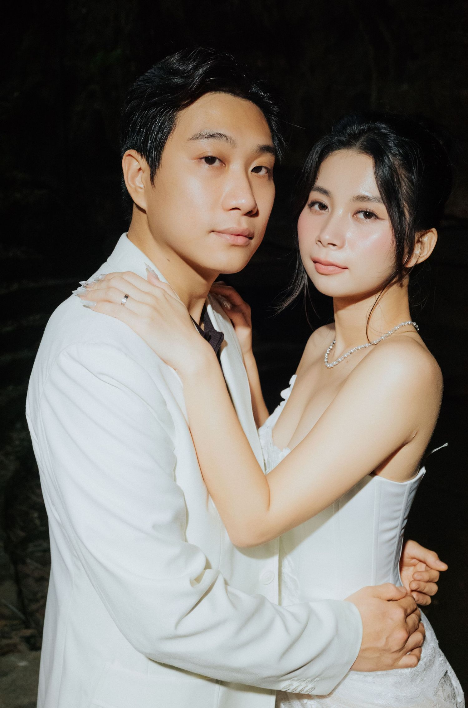 A couple in white outfits pose intimately against a dark background, the woman wearing a strapless dress and pearl necklace.