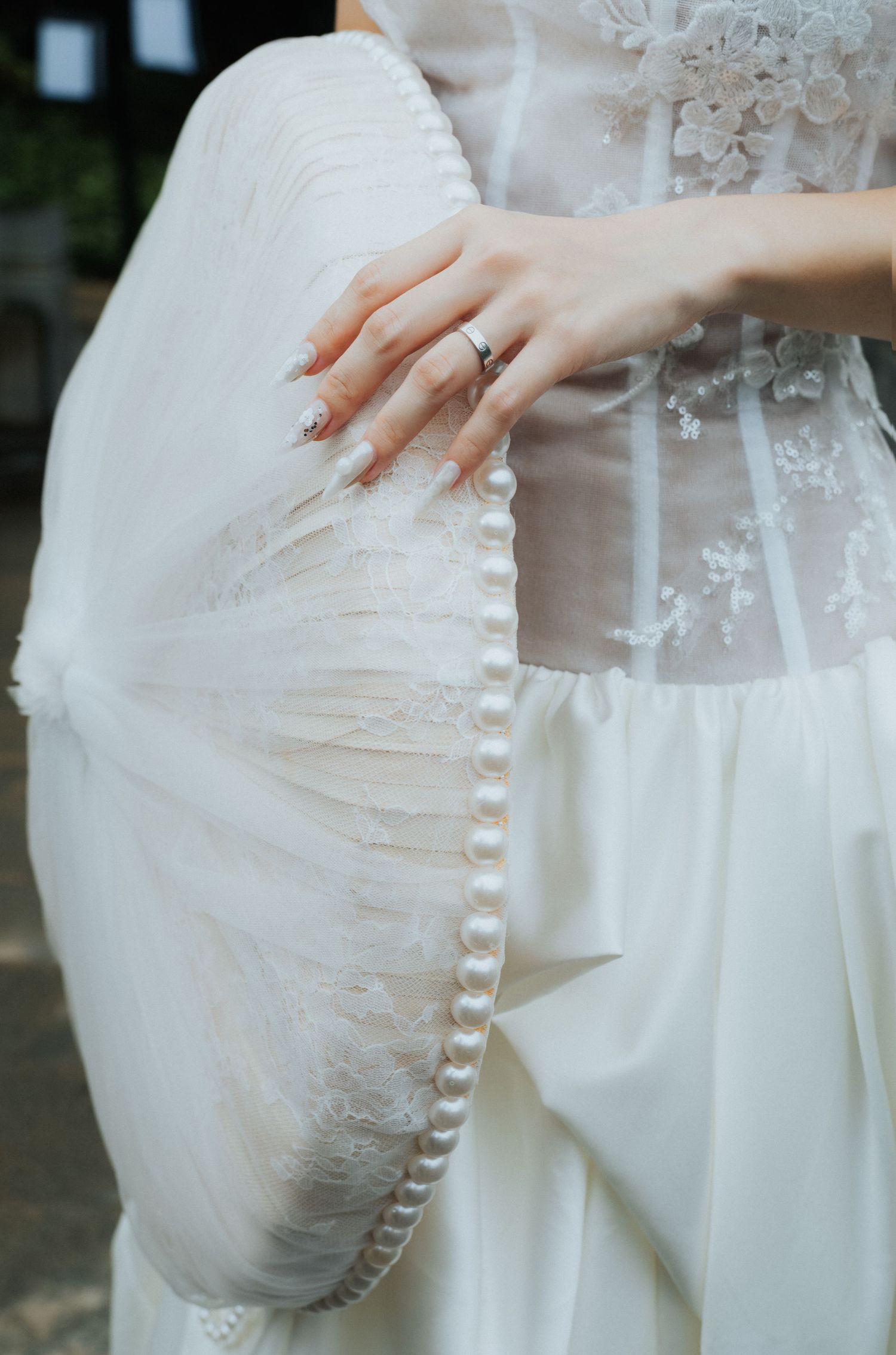 Bride in white lace wedding gown holding an elegant pearl-trimmed fan, wearing a delicate engagement ring.