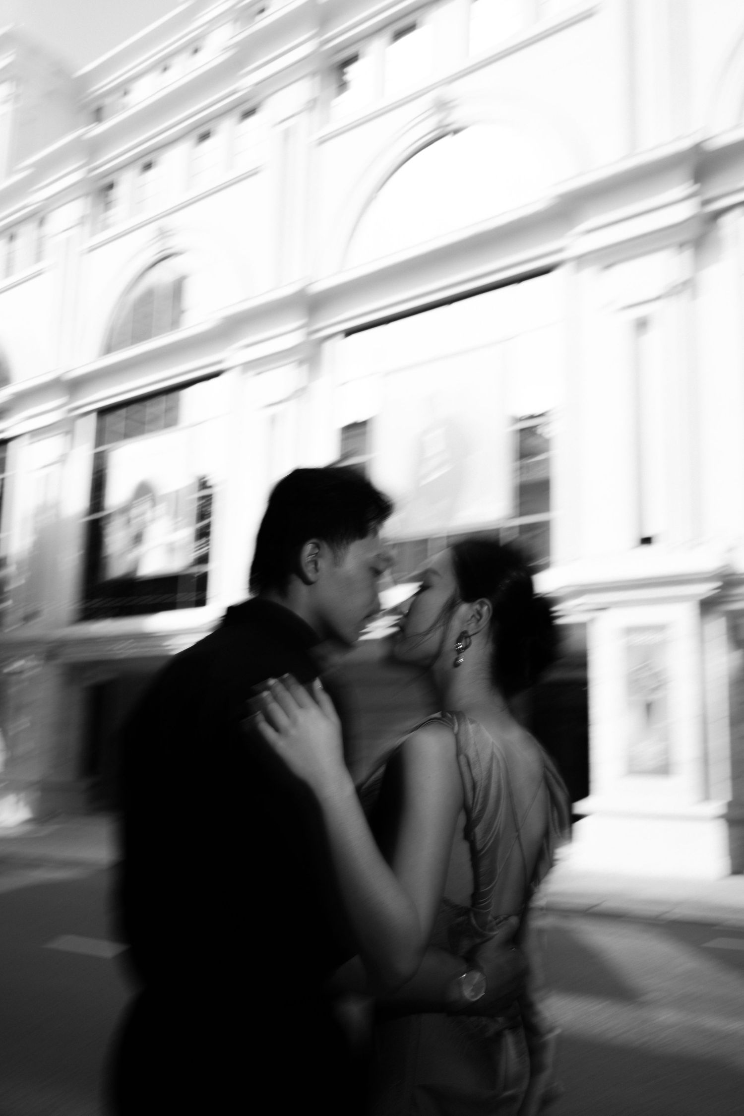 Black and white photo of a romantic couple embracing and kissing on a city street outside a grand building.