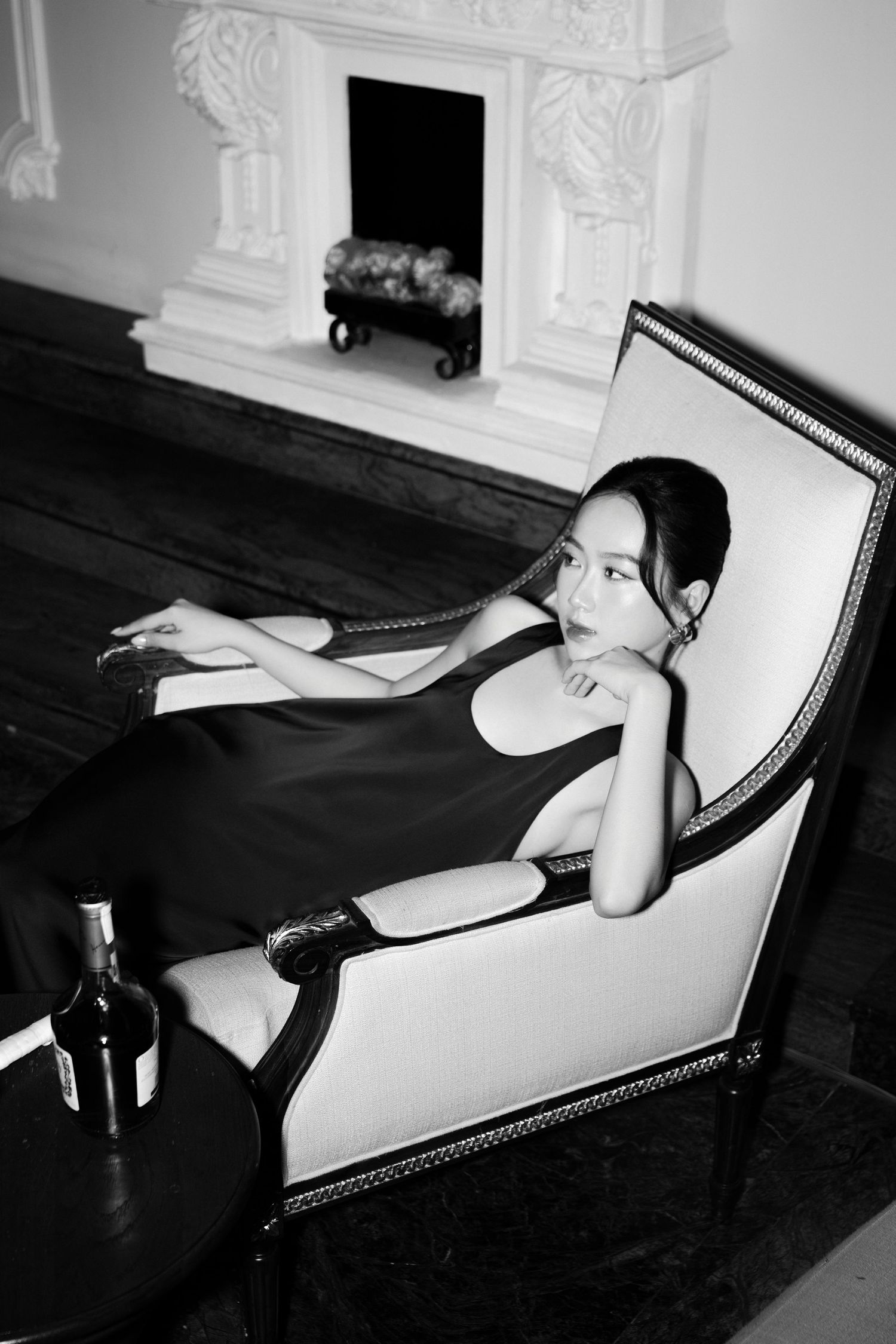 Black and white photo of elegant woman in black dress reclining in ornate chair near fireplace.
