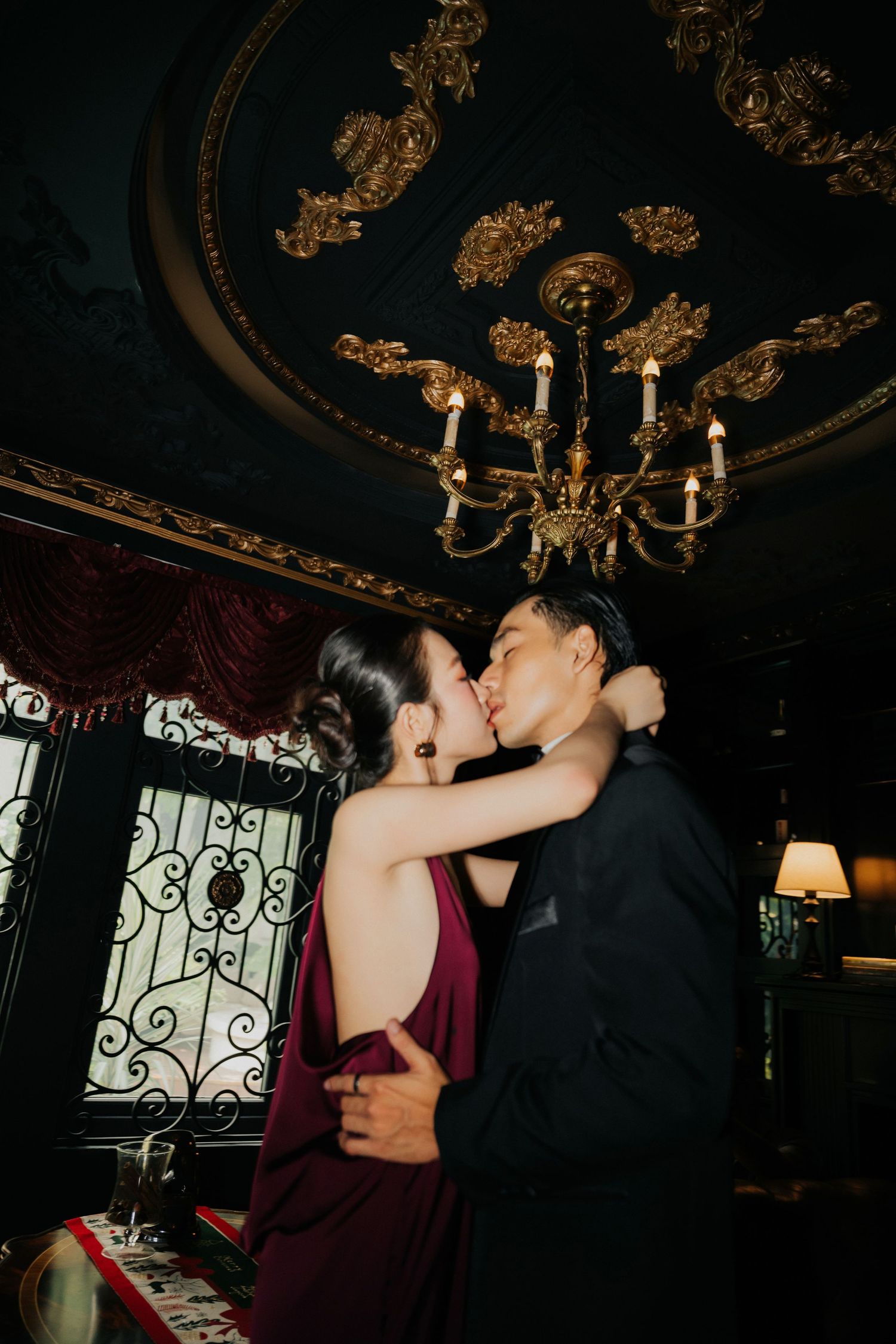 Couple sharing a romantic kiss in an elegant dark interior with ornate gold chandelier and ceiling details.