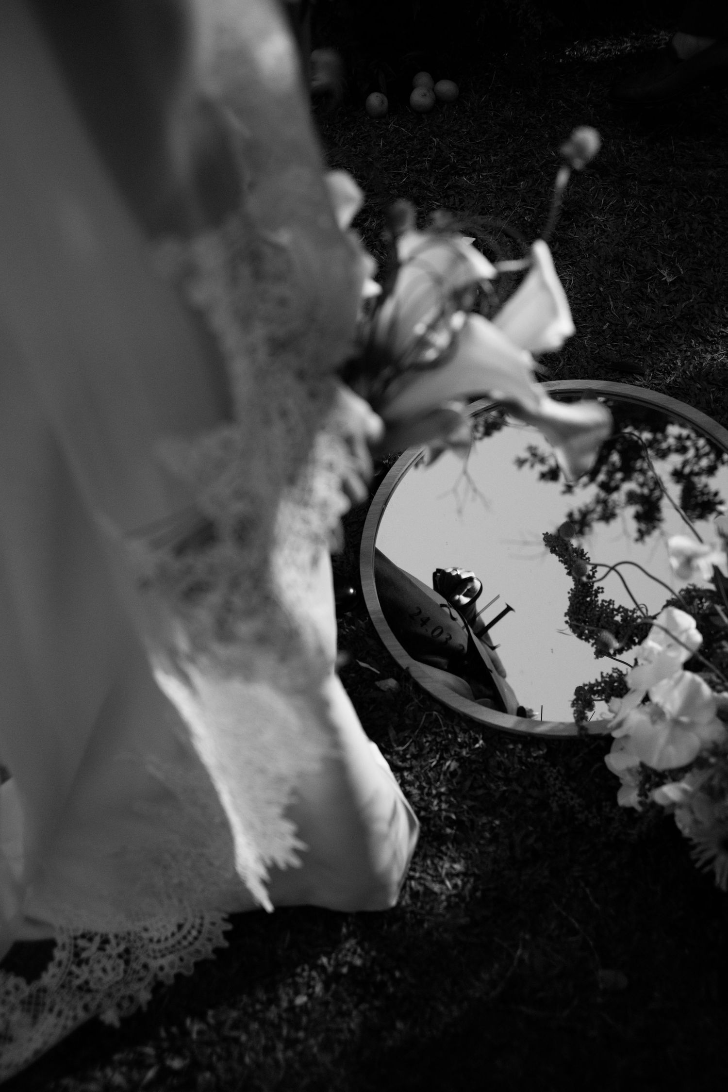 Black and white wedding photo with circular mirror on ground reflecting bouquet and bride in lace gown outdoors.
