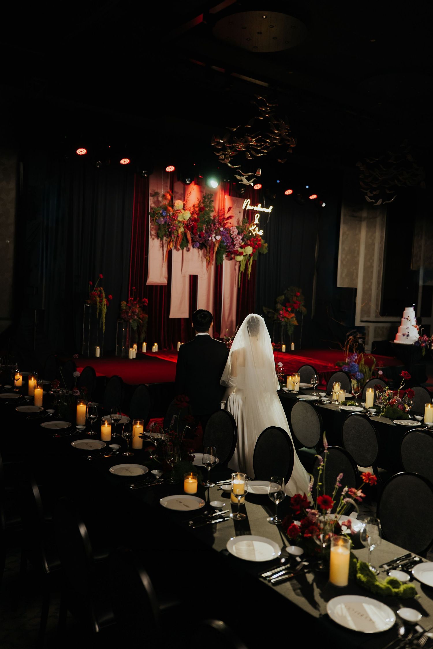 Bride and groom stand at a dark, moody wedding reception with black tables, candles, and vibrant floral backdrop.