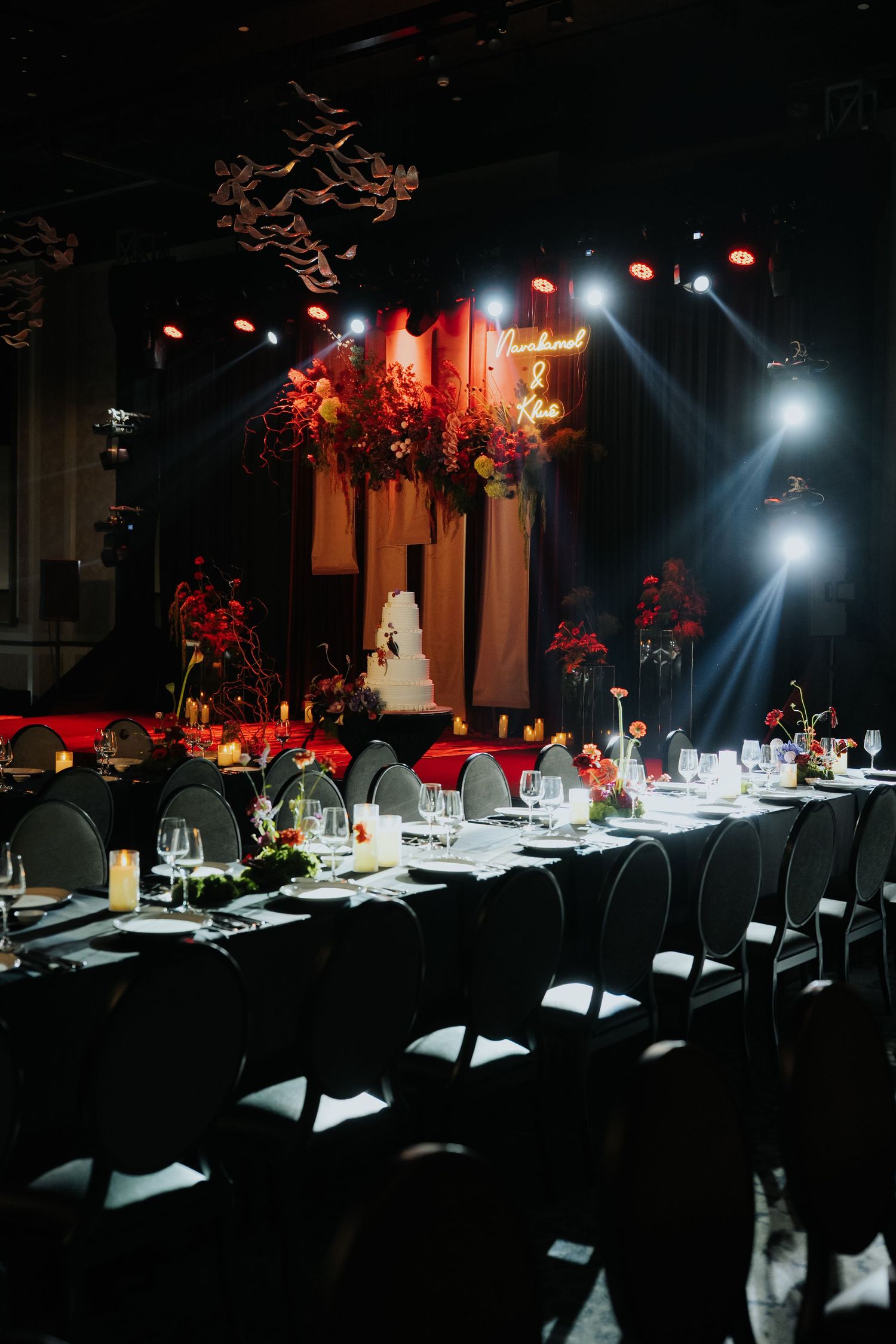 Elegant wedding reception with long black tables, floral arrangements, stage with white cake, neon sign, and dramatic red lighting.