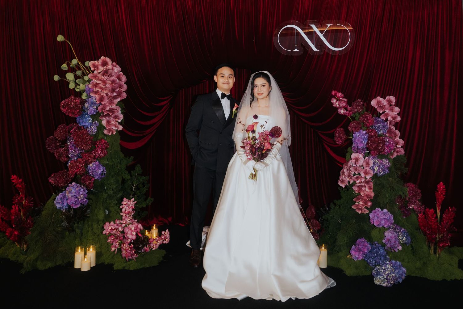 Elegant wedding couple posing before a red floral backdrop with orchids and greenery in a formal venue.