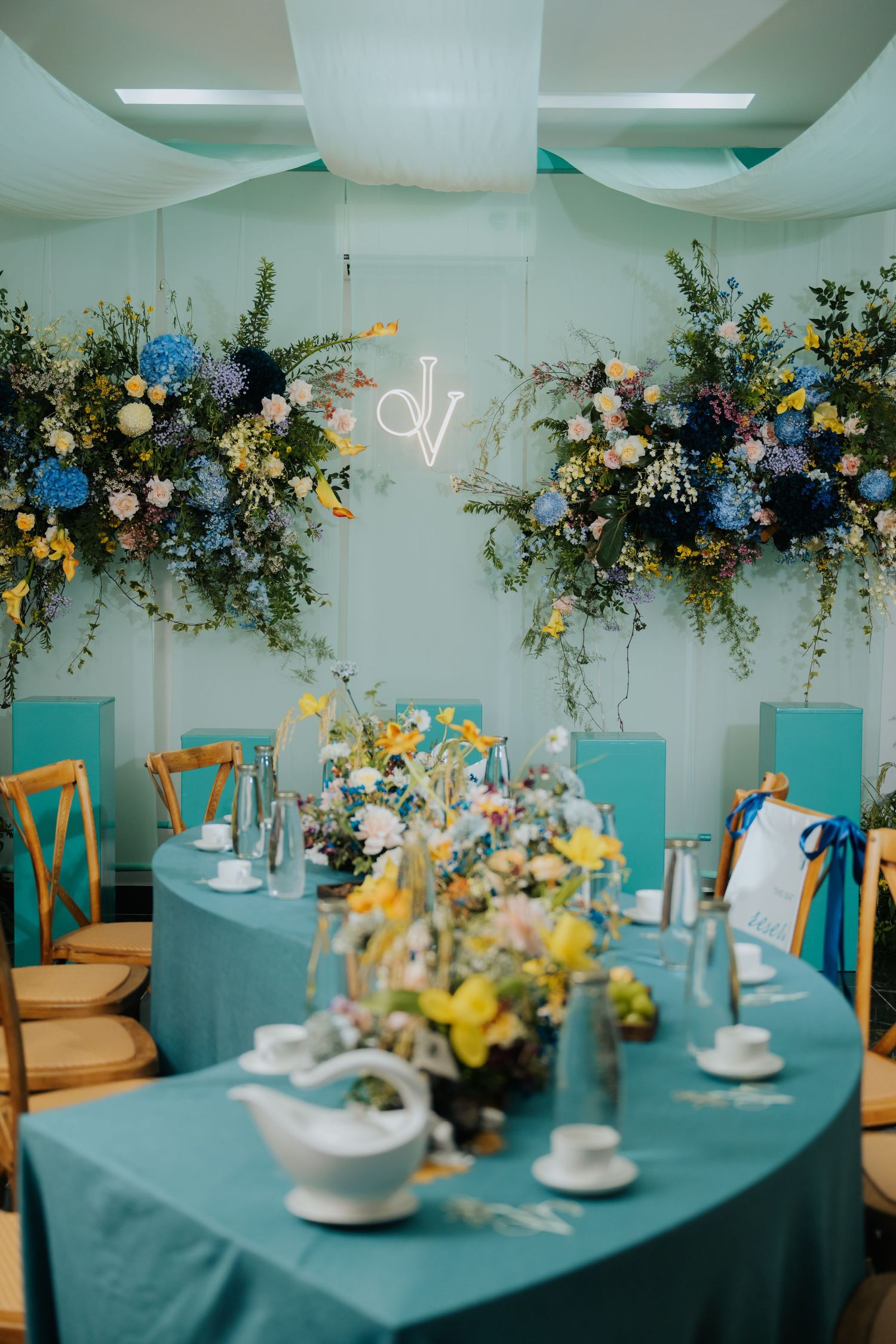 Elegant wedding reception with teal tablecloths, floral arrangements, wooden chairs, and a neon monogram sign on a blue wall.