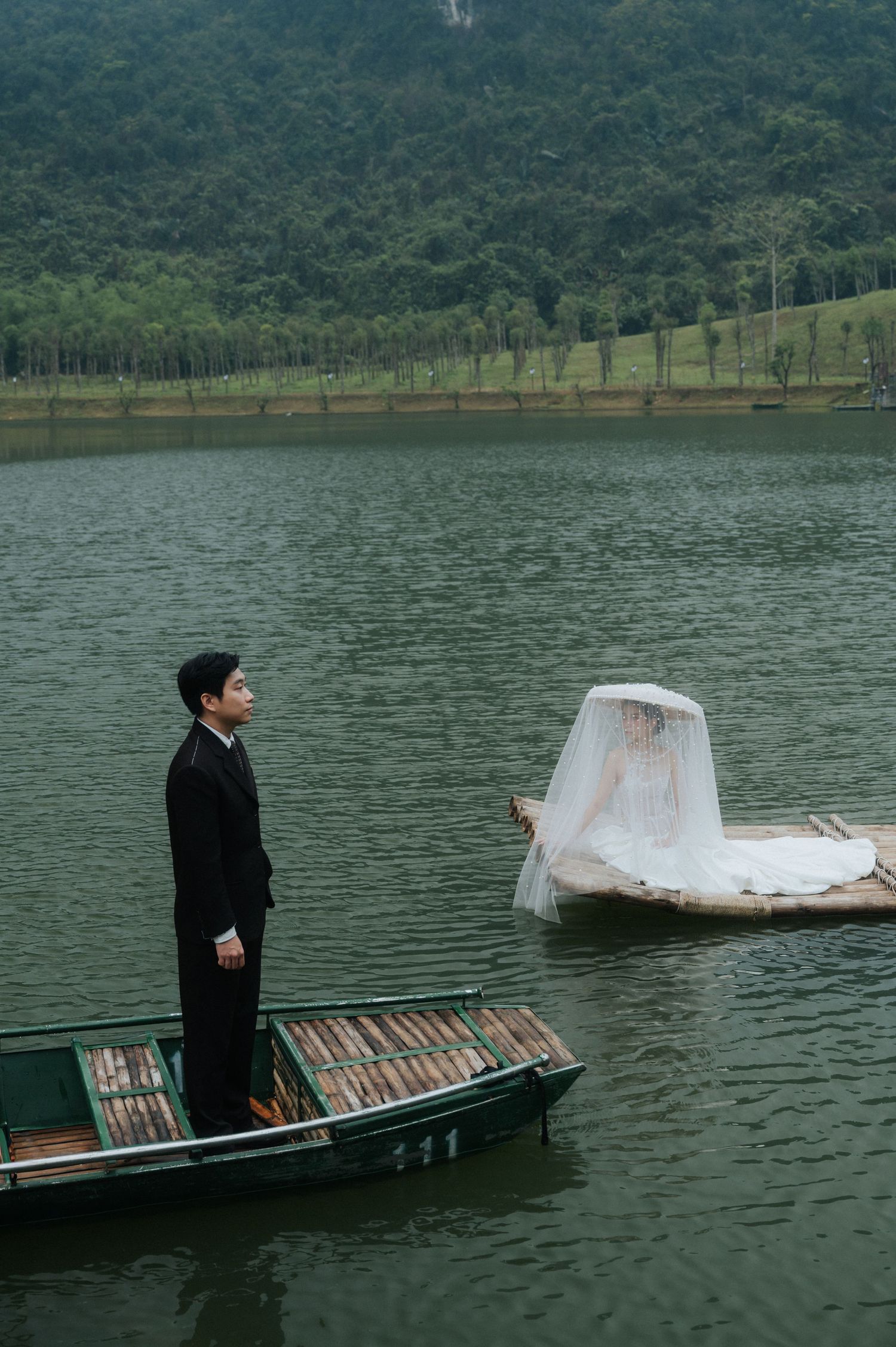 Groom in black suit stands on wooden boat facing bride in white wedding dress on separate boat on a serene lake.
