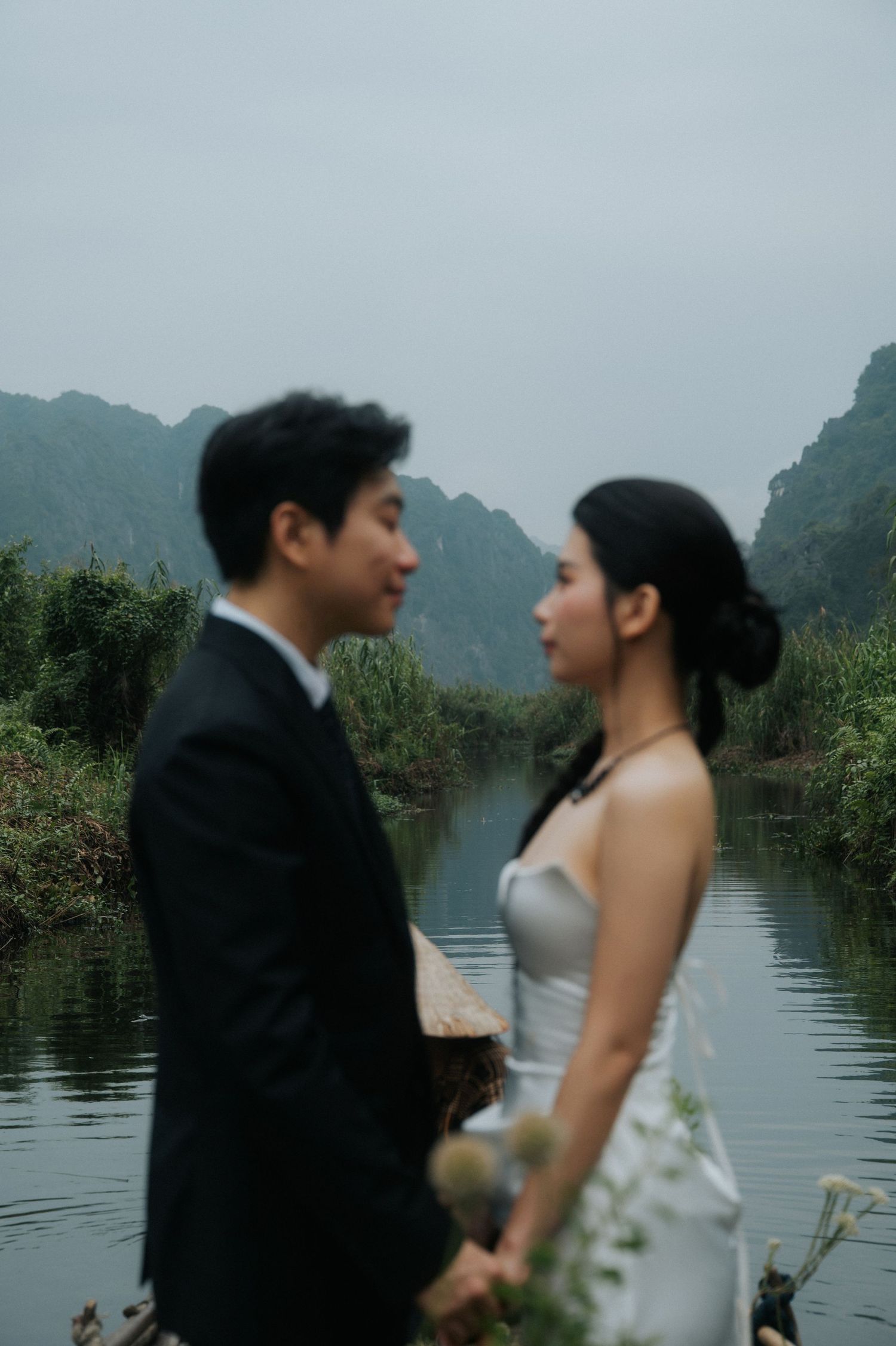Couple in wedding attire sharing a romantic moment by a misty river with karst mountains in the background.