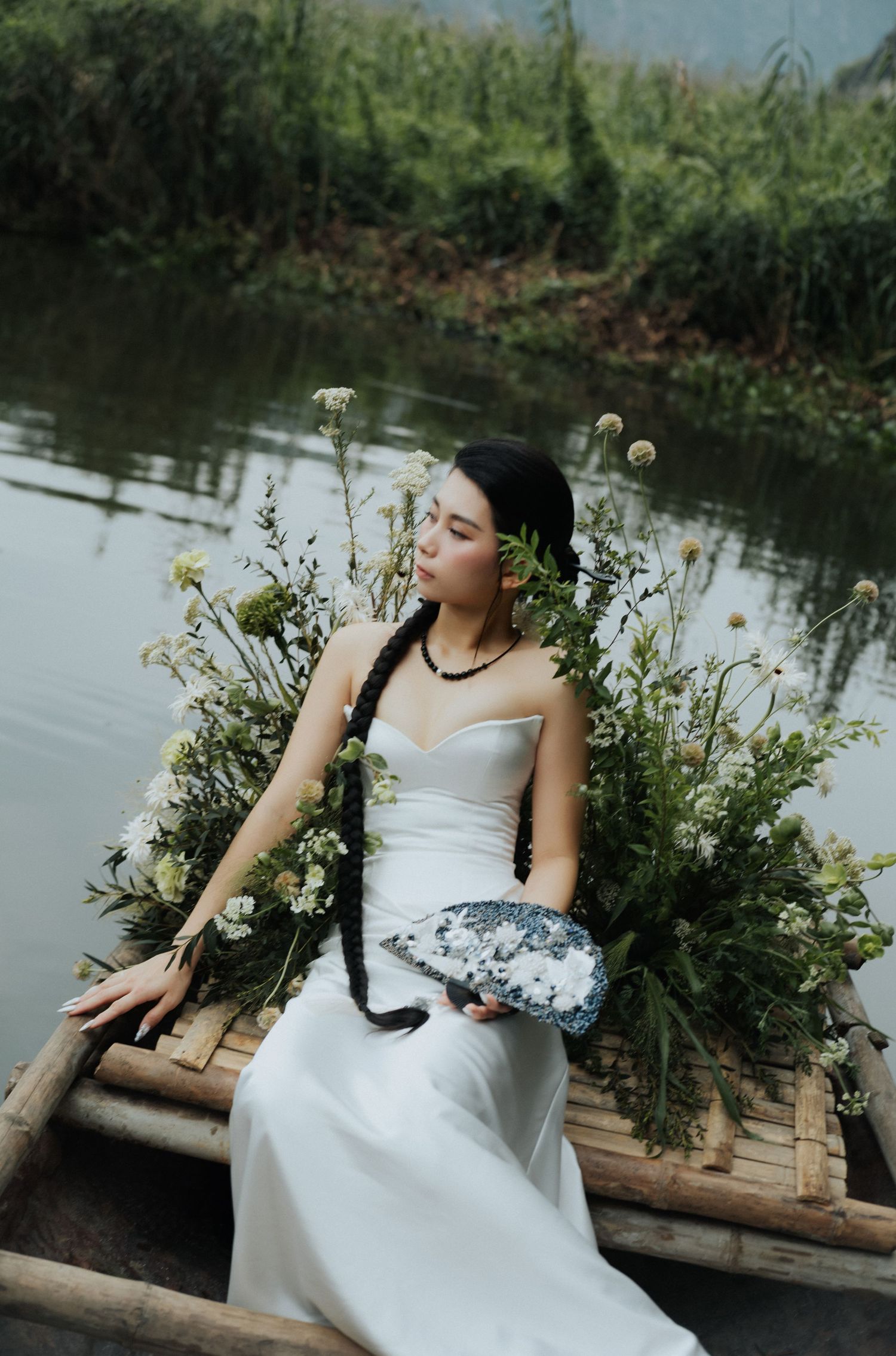 Woman in white gown sits on bamboo boat holding blue crystal, surrounded by wildflowers on a serene river.