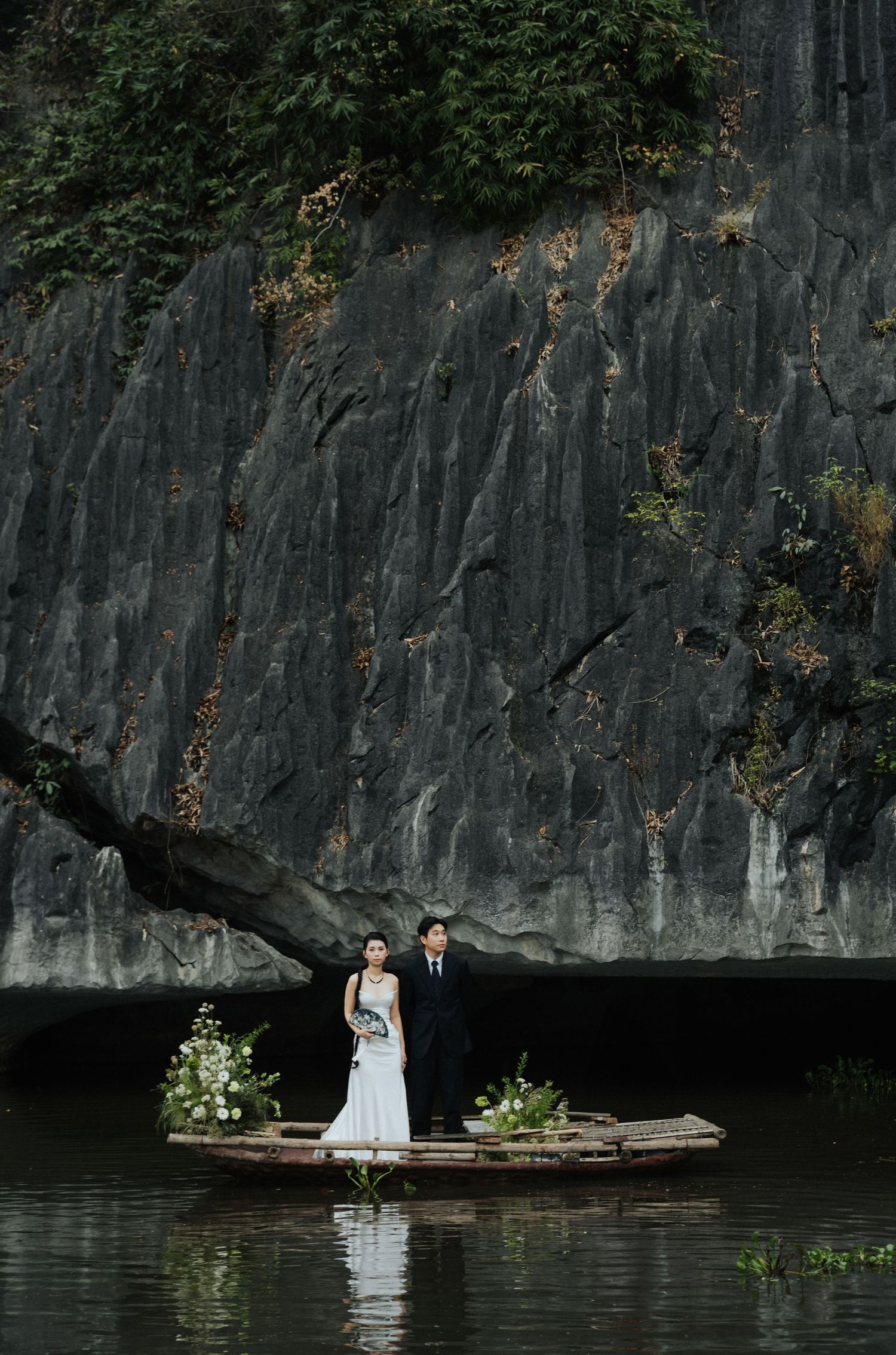 Bride and groom stand on a wooden boat with floral arrangements before a dramatic dark rock cliff in Ninh Binh.