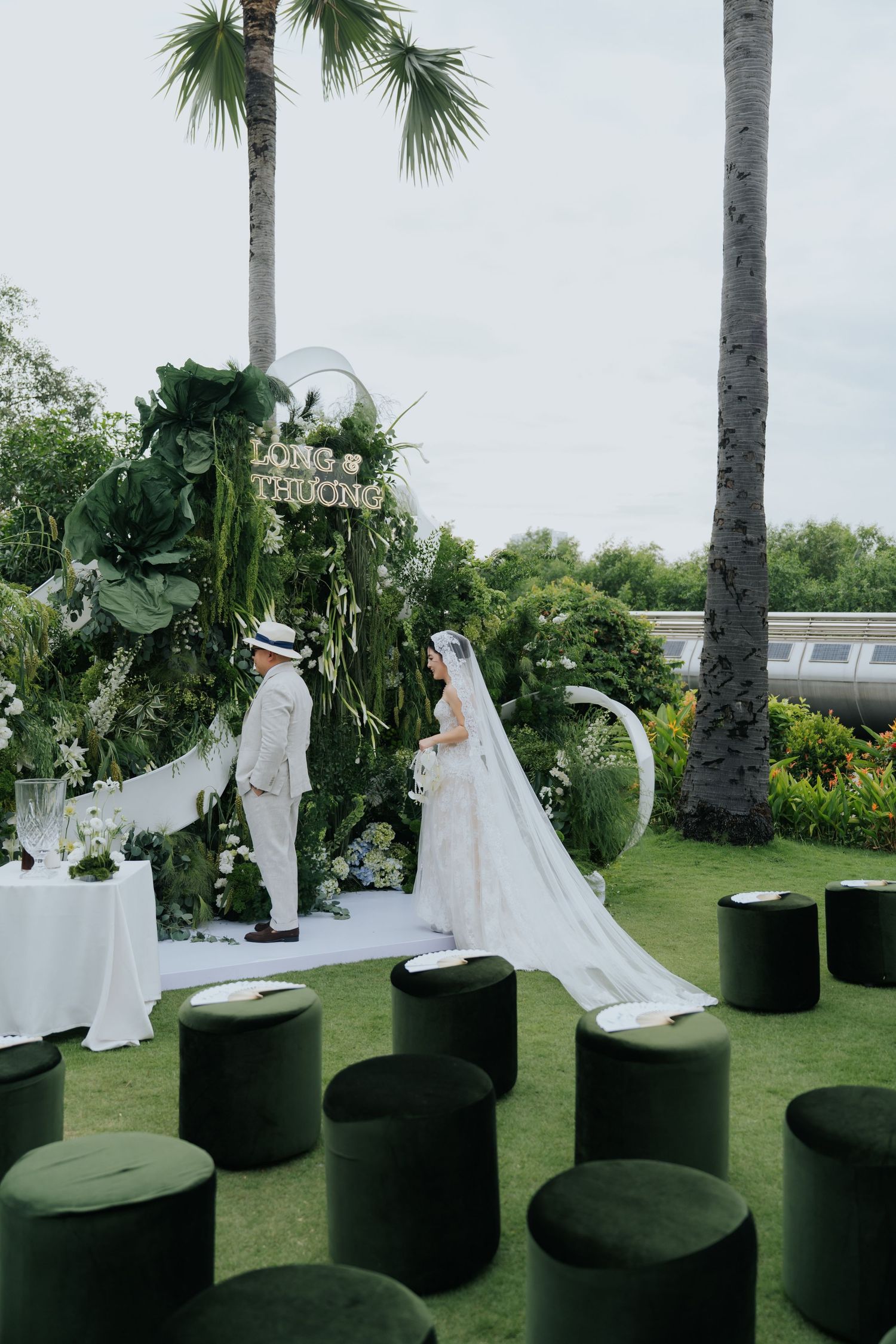 Outdoor wedding ceremony on green lawn with couple at floral arch, palm trees, and elegant green velvet seating.