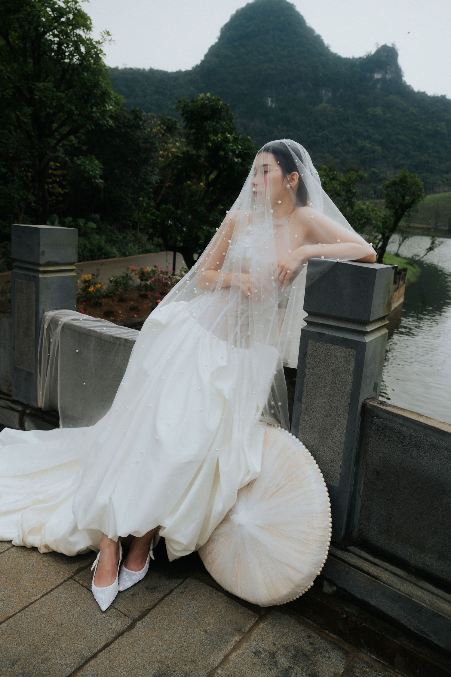 Bride in white gown and veil poses by stone railing with karst mountains and lake in background.